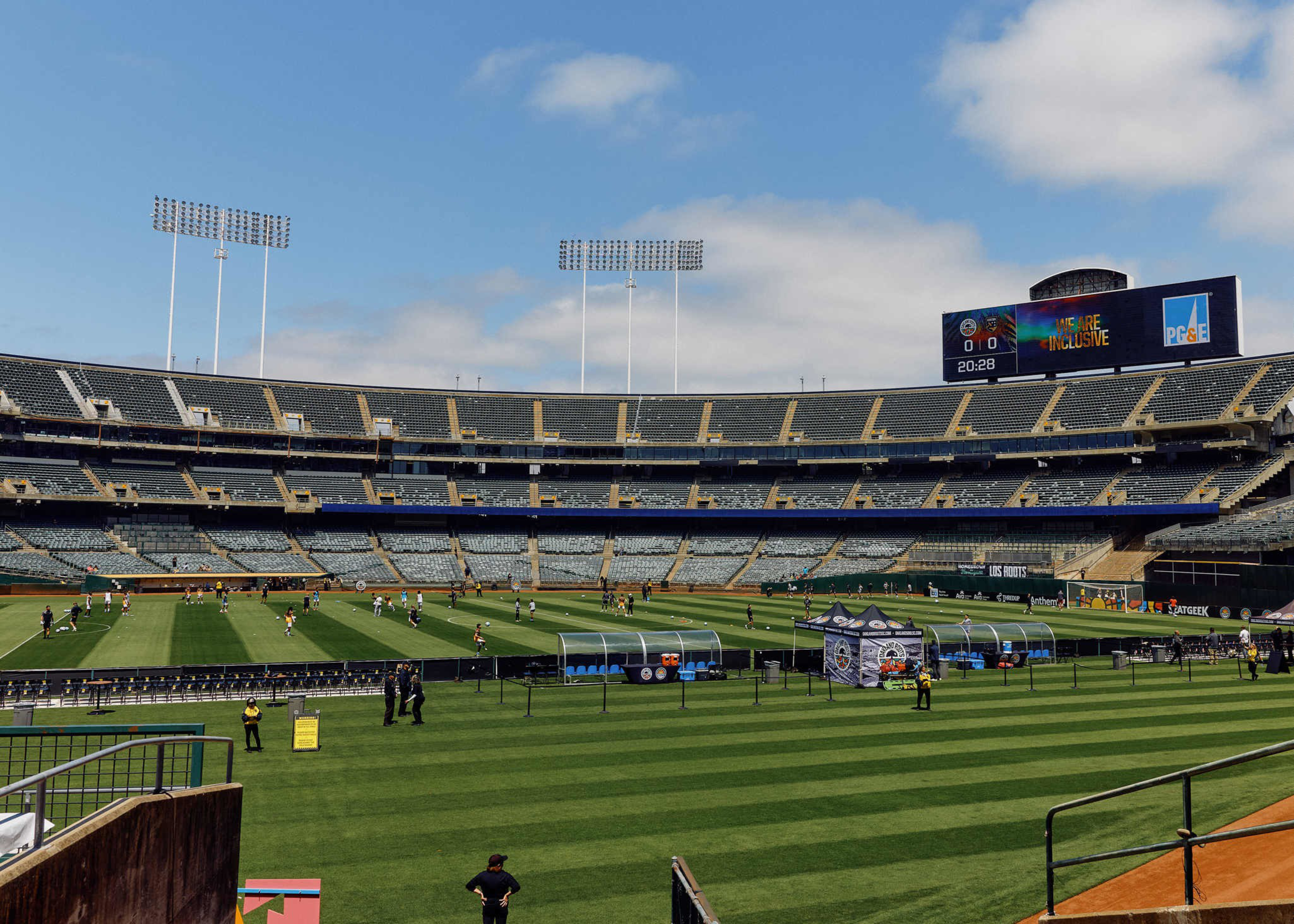 A nearly empty stadium with players warming up on the field under a partly cloudy sky and a scoreboard displaying a message about inclusivity.