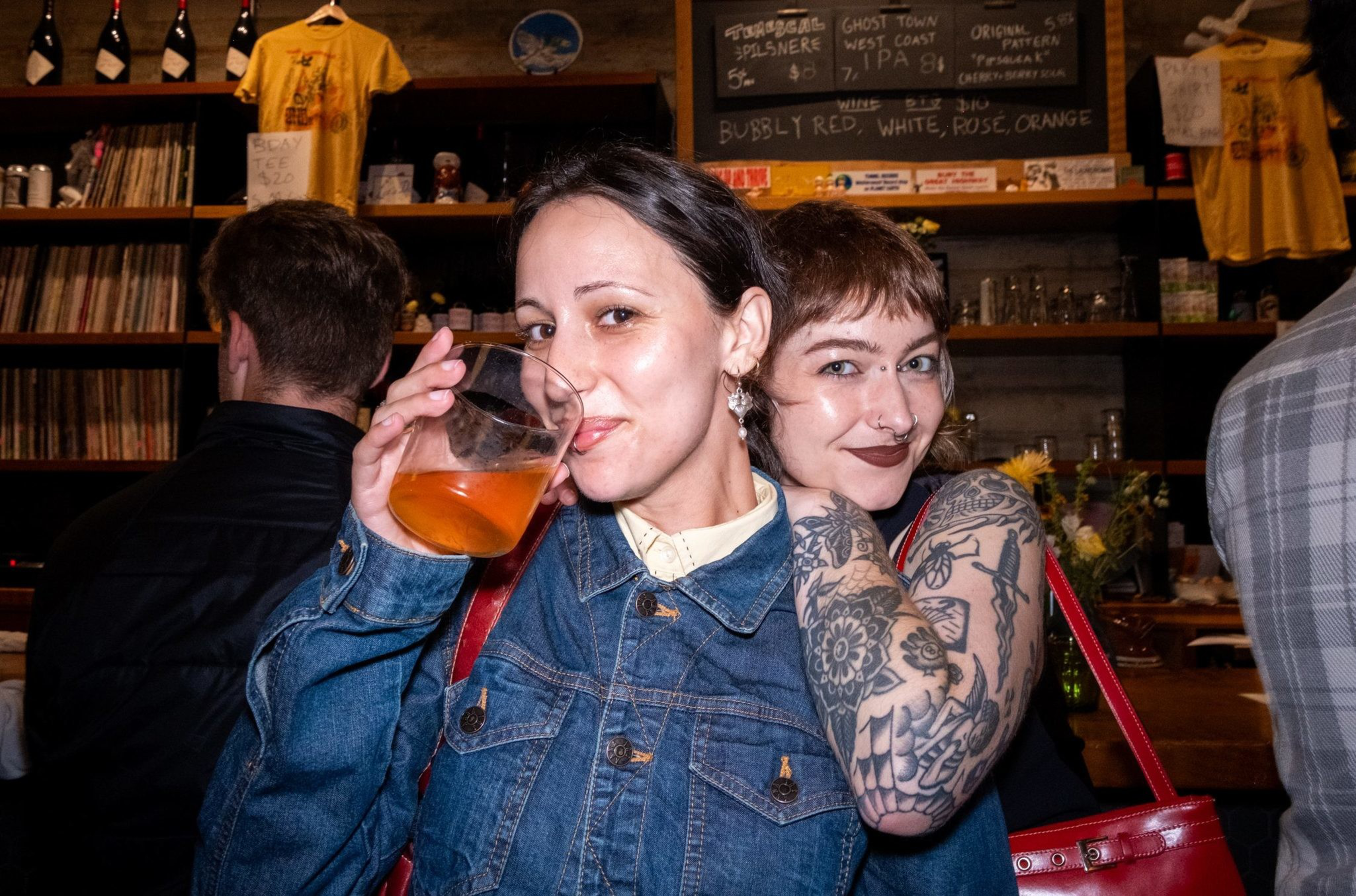 Two smiling women stand in a bar, one holding a drink and wearing a denim jacket, the other with tattoos and a red purse. Shelves and a menu are visible behind them.