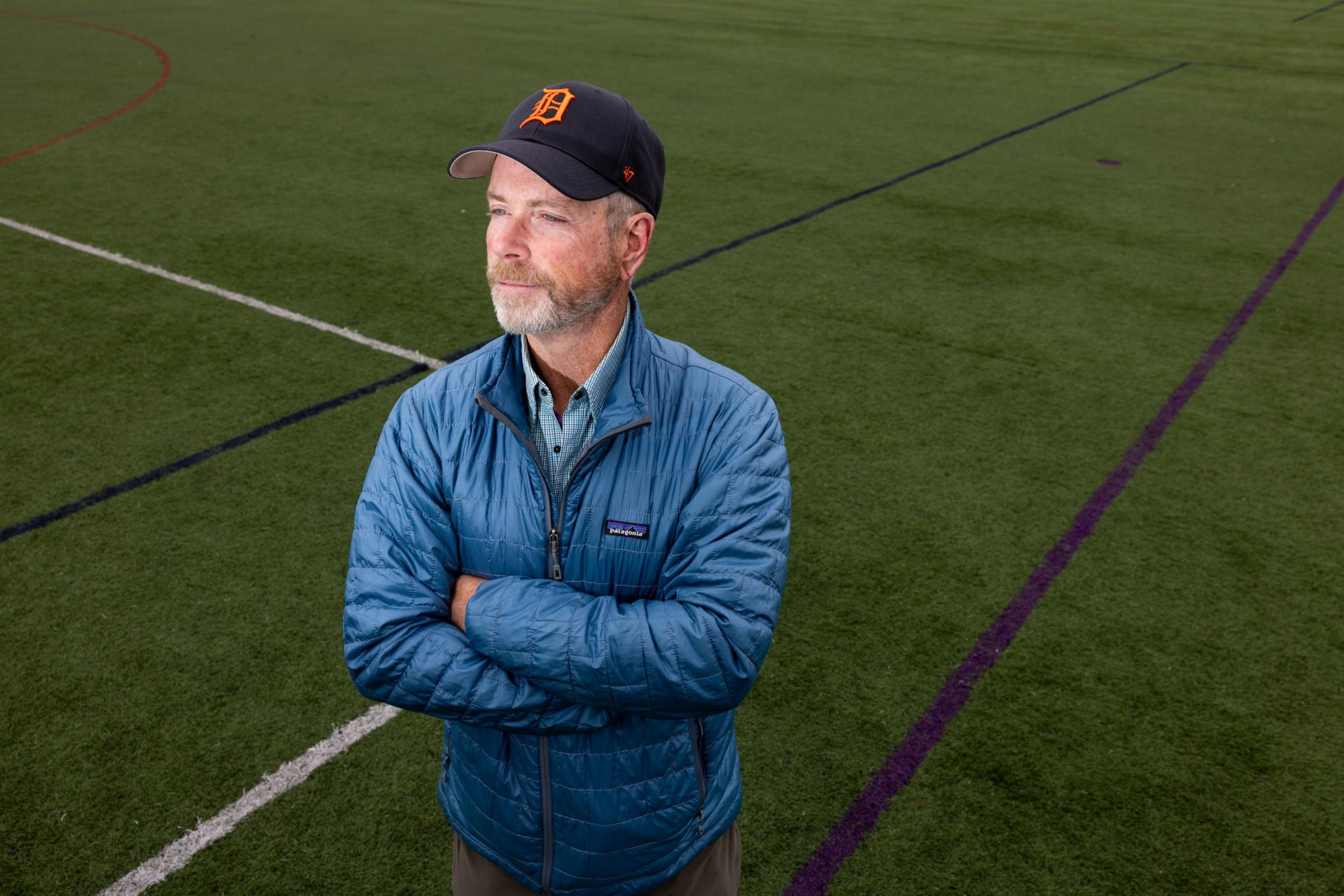 A man stands on a sports field with crossed arms, wearing a blue jacket and a black cap. The field has various colored lines on the green grass.