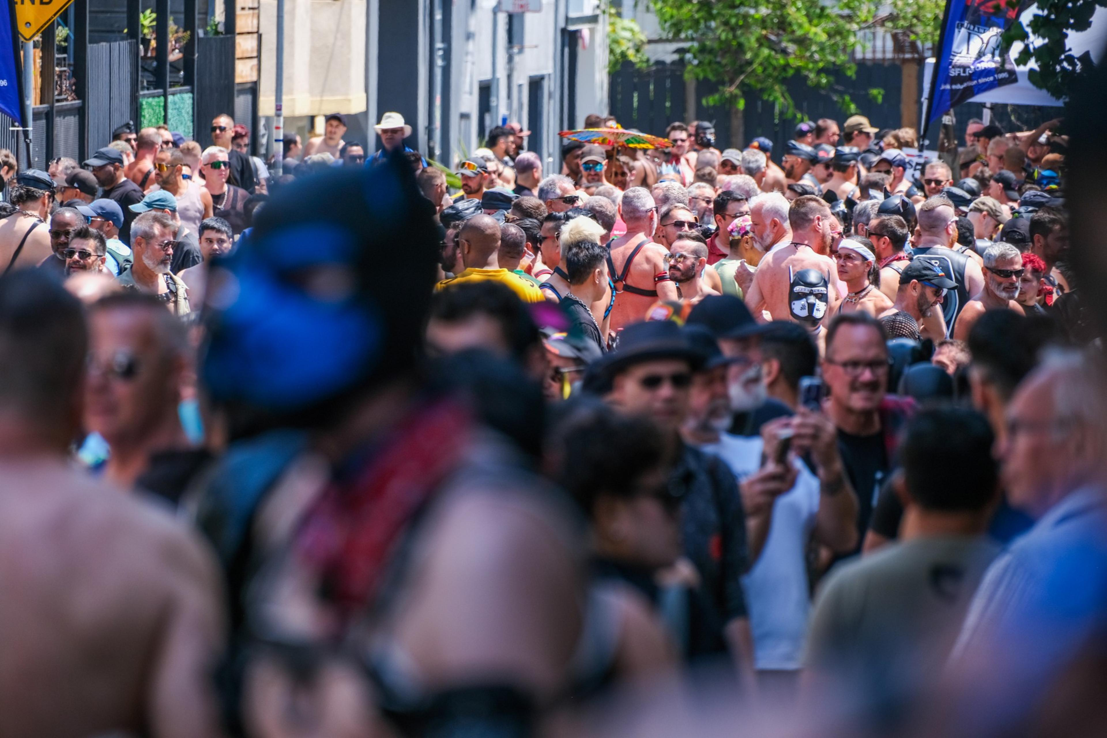 A large crowd of people fills a street, many dressed in leather and unique costumes. It's a lively, colorful scene, reflecting a festive atmosphere.