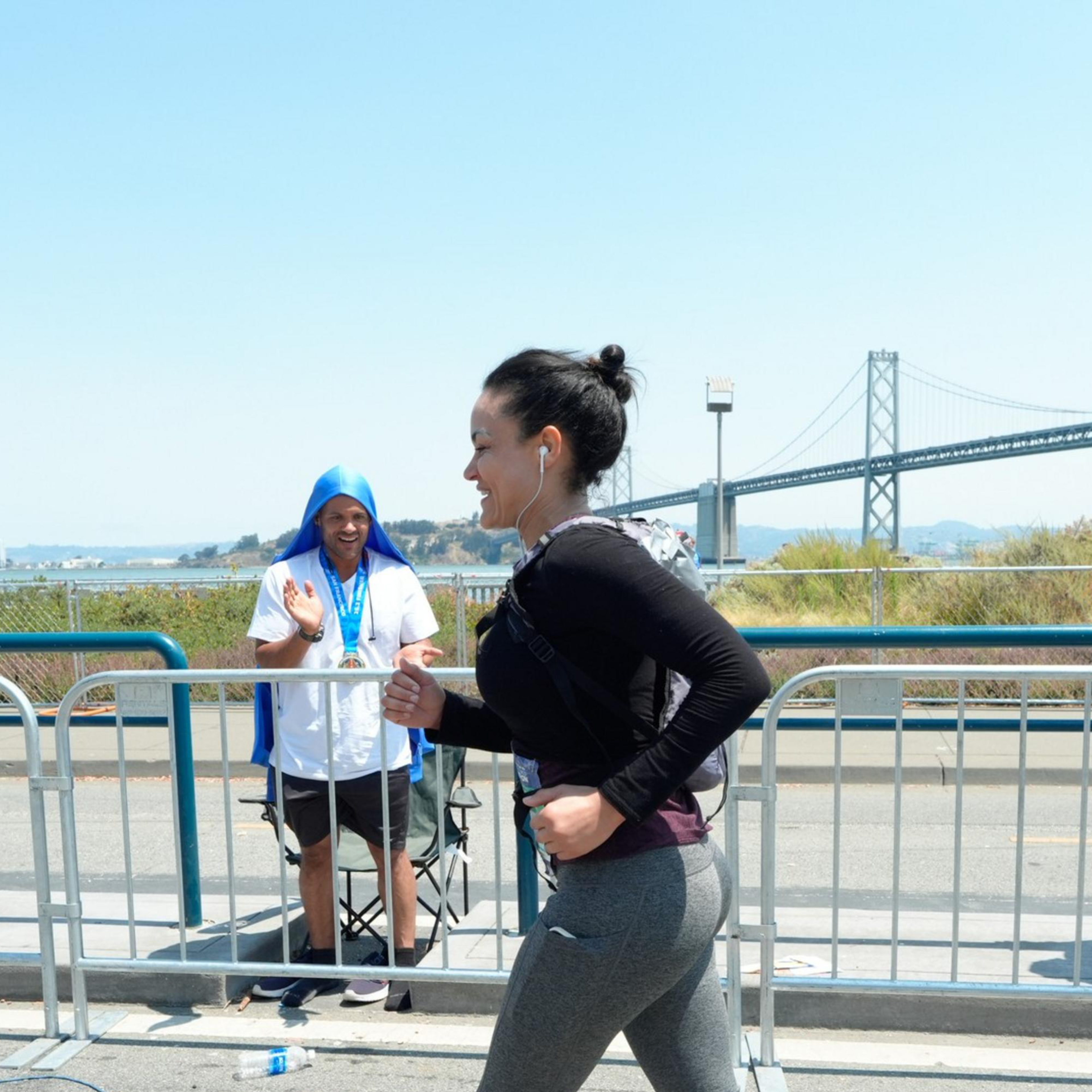 A woman jogs in a race with earbuds on, while two men with towels over their heads cheer her on behind a barrier near a bridge on a sunny day.