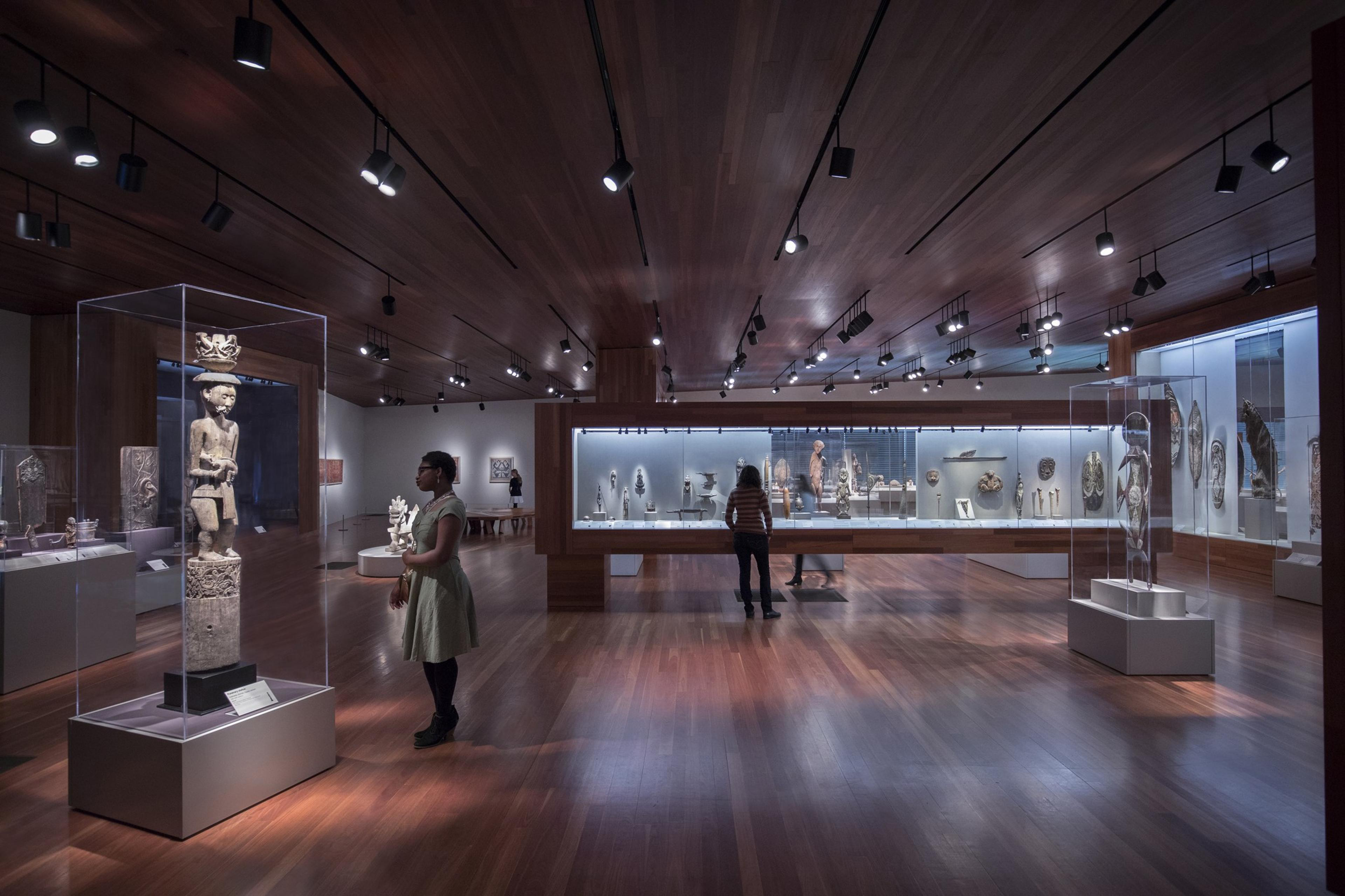 The image shows a museum gallery with wooden floors and ceiling, displaying various sculptures and artifacts in glass cases. Several people are observing the exhibits.
