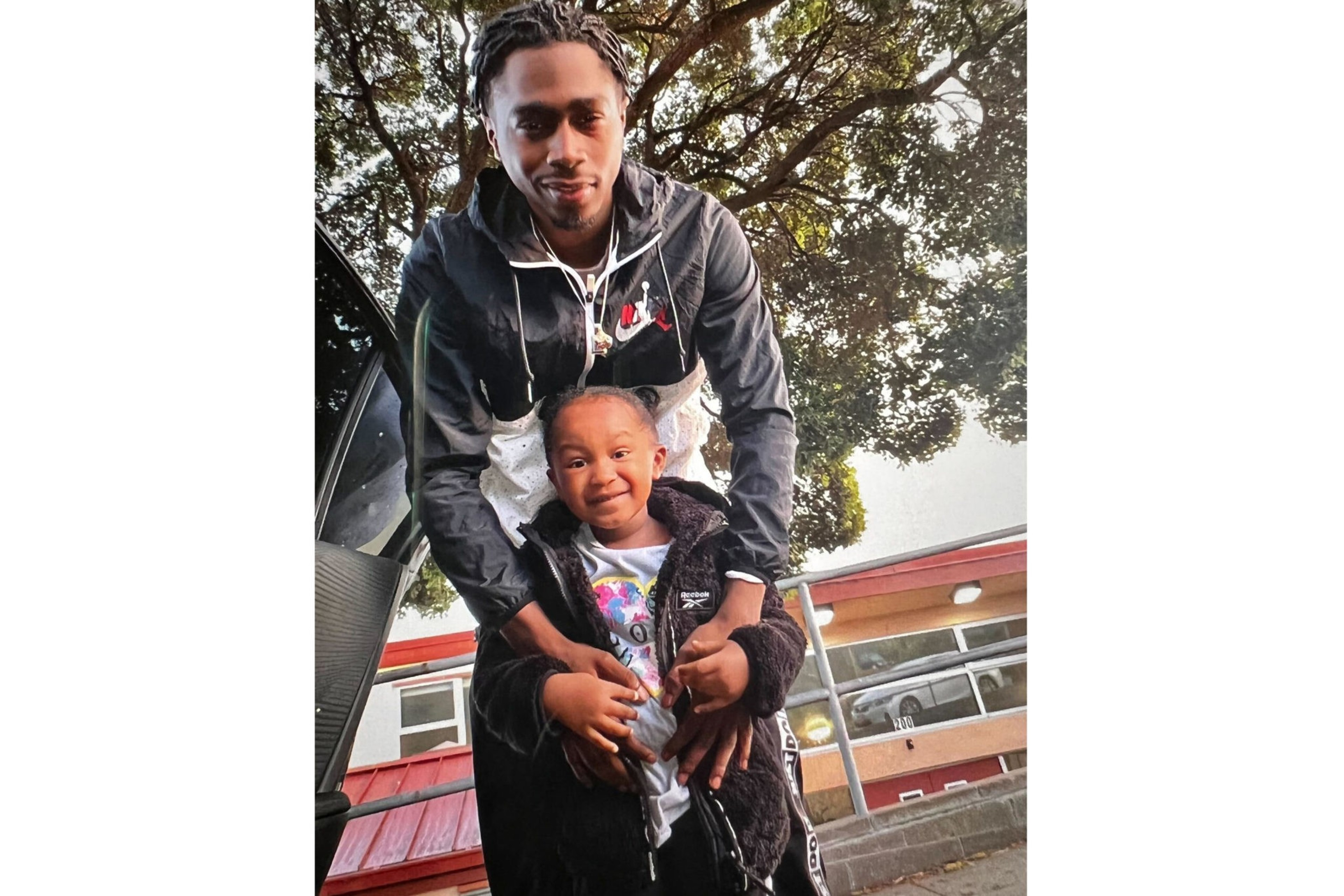 A man with braided hair gently holds a smiling young child dressed in a black jacket, standing near a car and a tree outdoors.