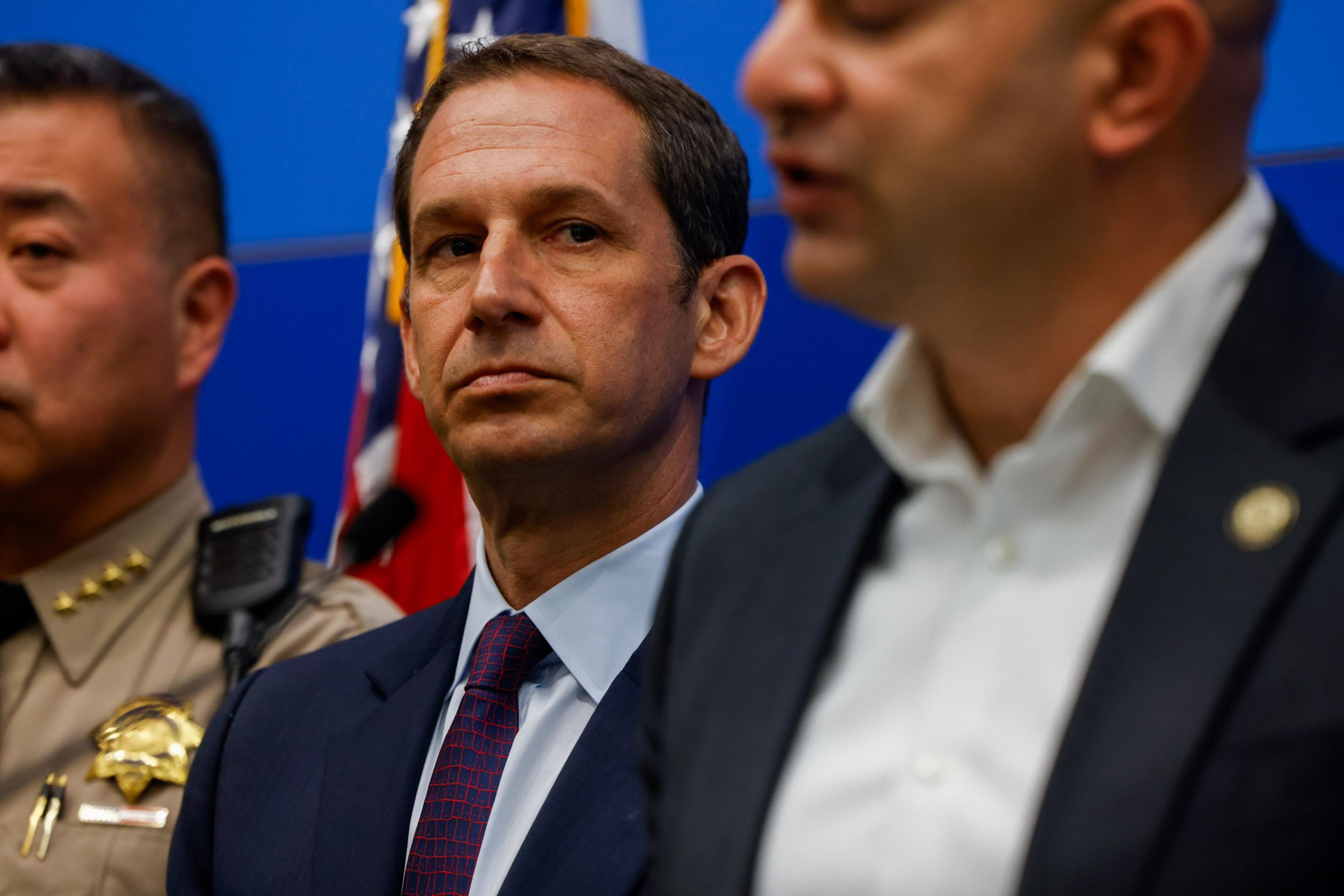 Three men in suits and uniforms are standing in a row, one focused, others partially out of frame, against a blue background with flags.