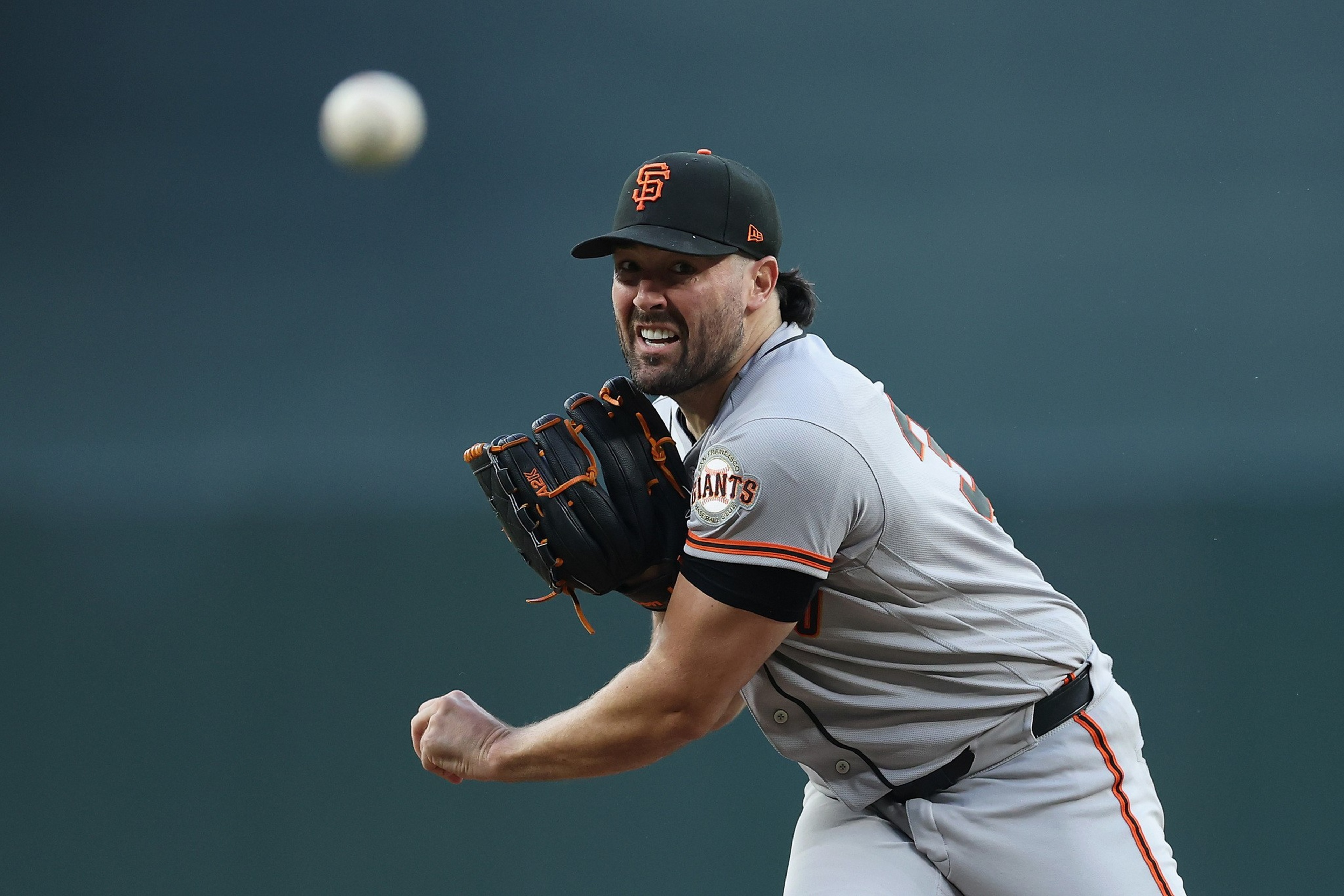 A baseball pitcher in a gray San Francisco Giants uniform is in mid-throw, with intense focus on his face and the ball visible in the air.