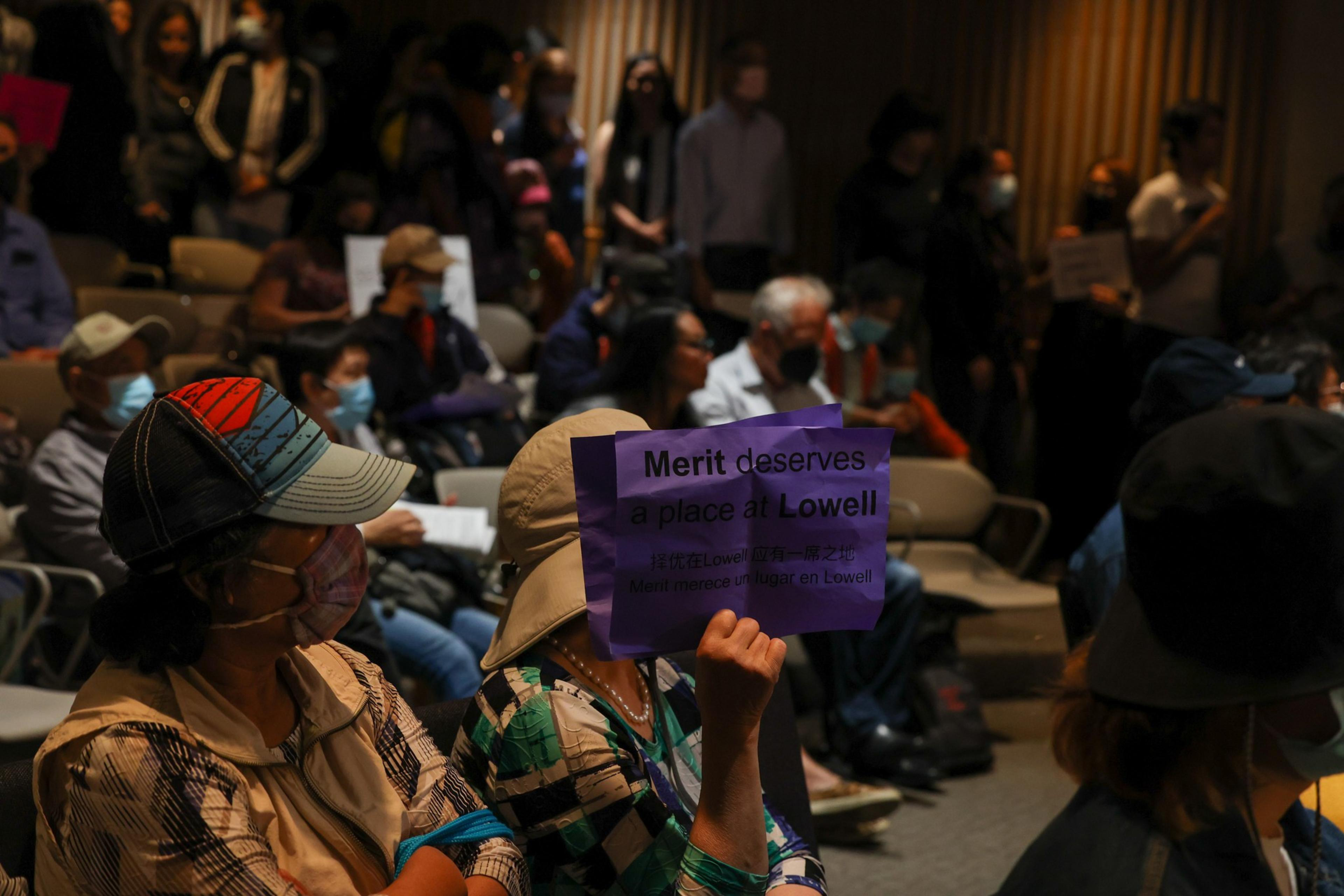 People wearing masks sit and stand closely together indoors, one person in the foreground holds a purple sign that reads, "Merit deserves a place at Lowell."