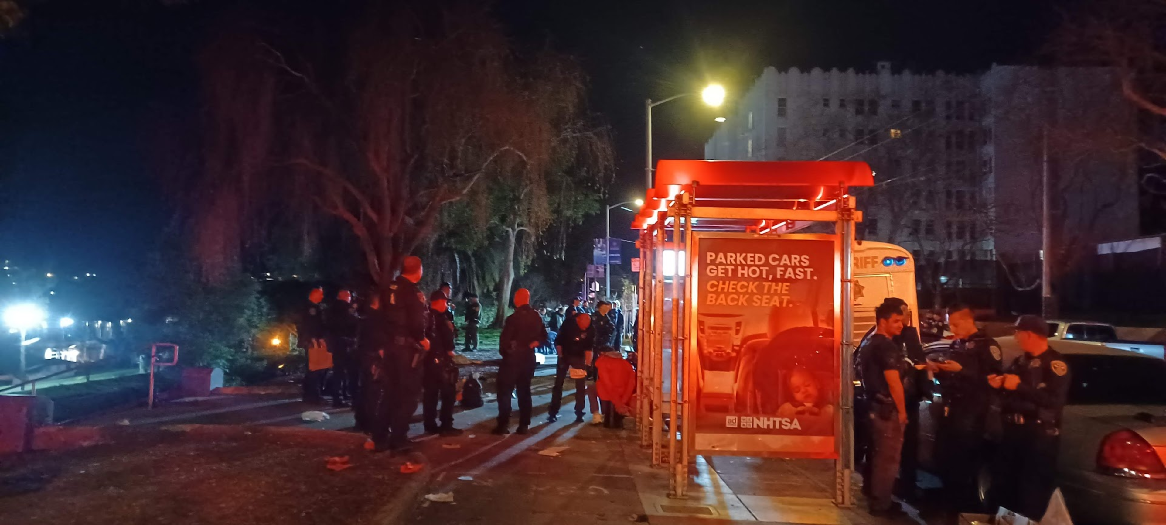 A nighttime scene with a group of police officers standing near a brightly lit bus stop. The bus stop displays an ad about parked cars getting hot quickly.