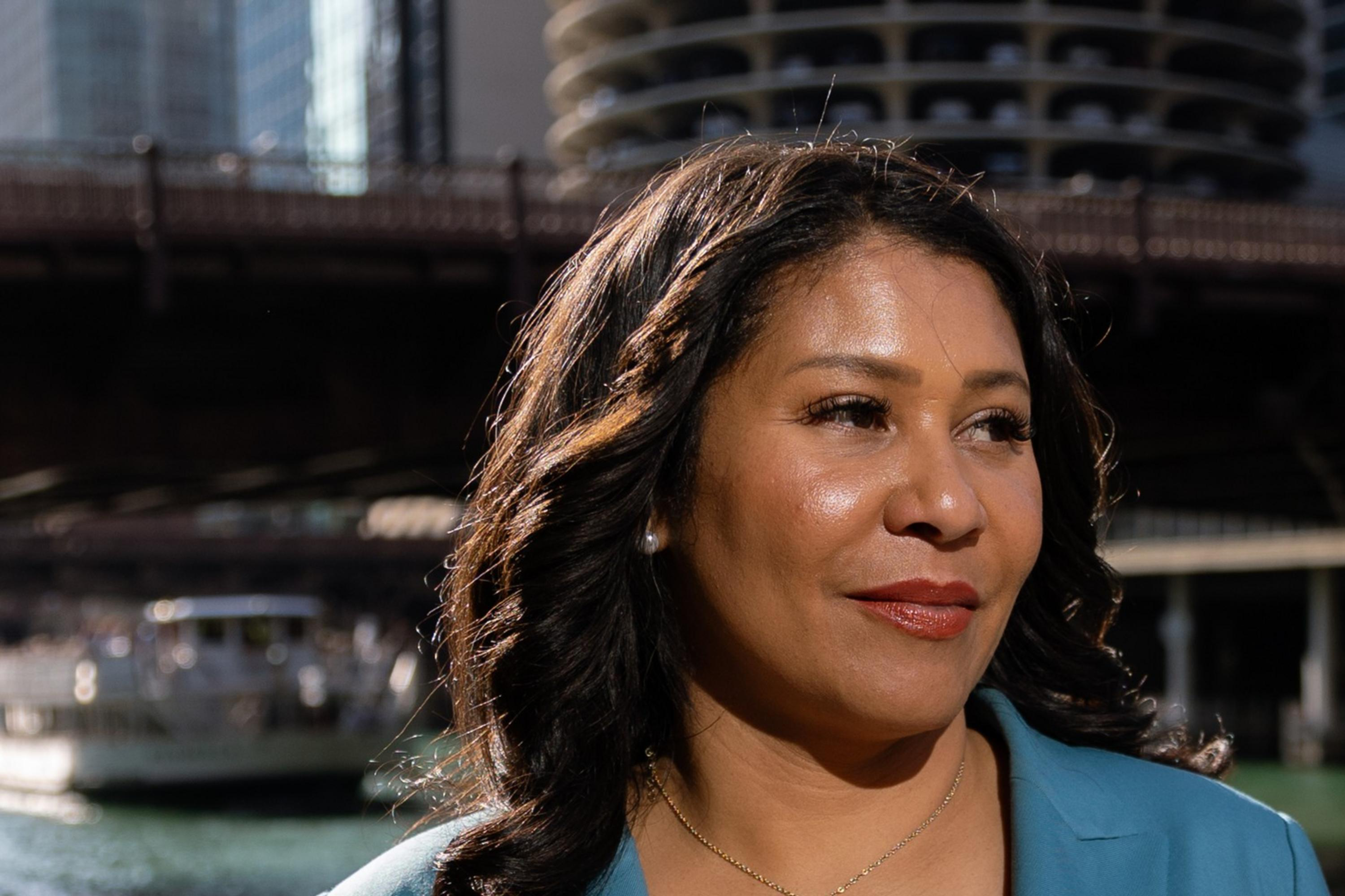 A woman in a blue blazer stands near a river with city buildings in the background. She has a circular pin on her lapel and appears thoughtful.