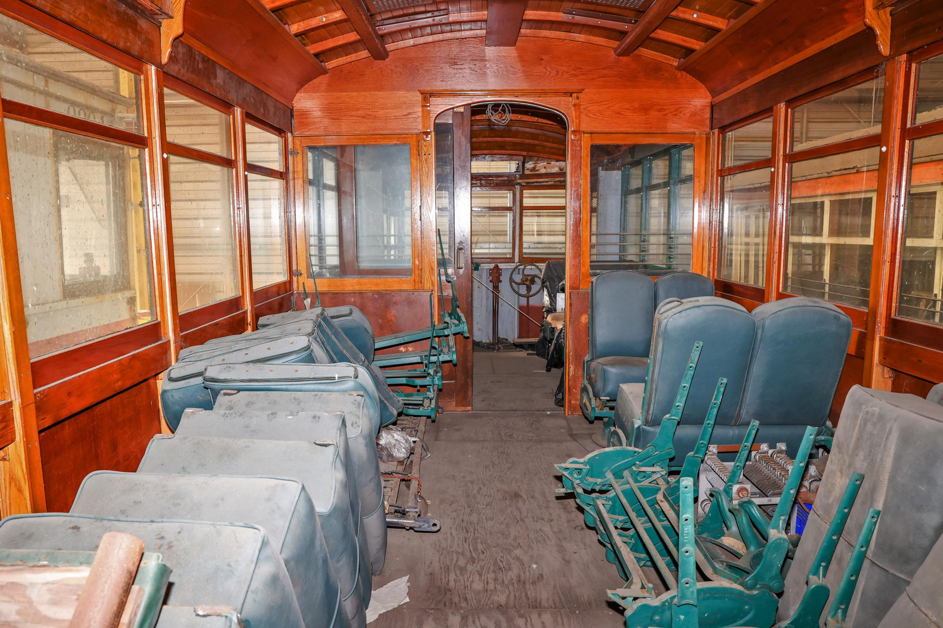 Old wooden trolley interior with worn, dusty blue and green seats, large windows, and a view toward the driver’s cabin with a steering wheel.
