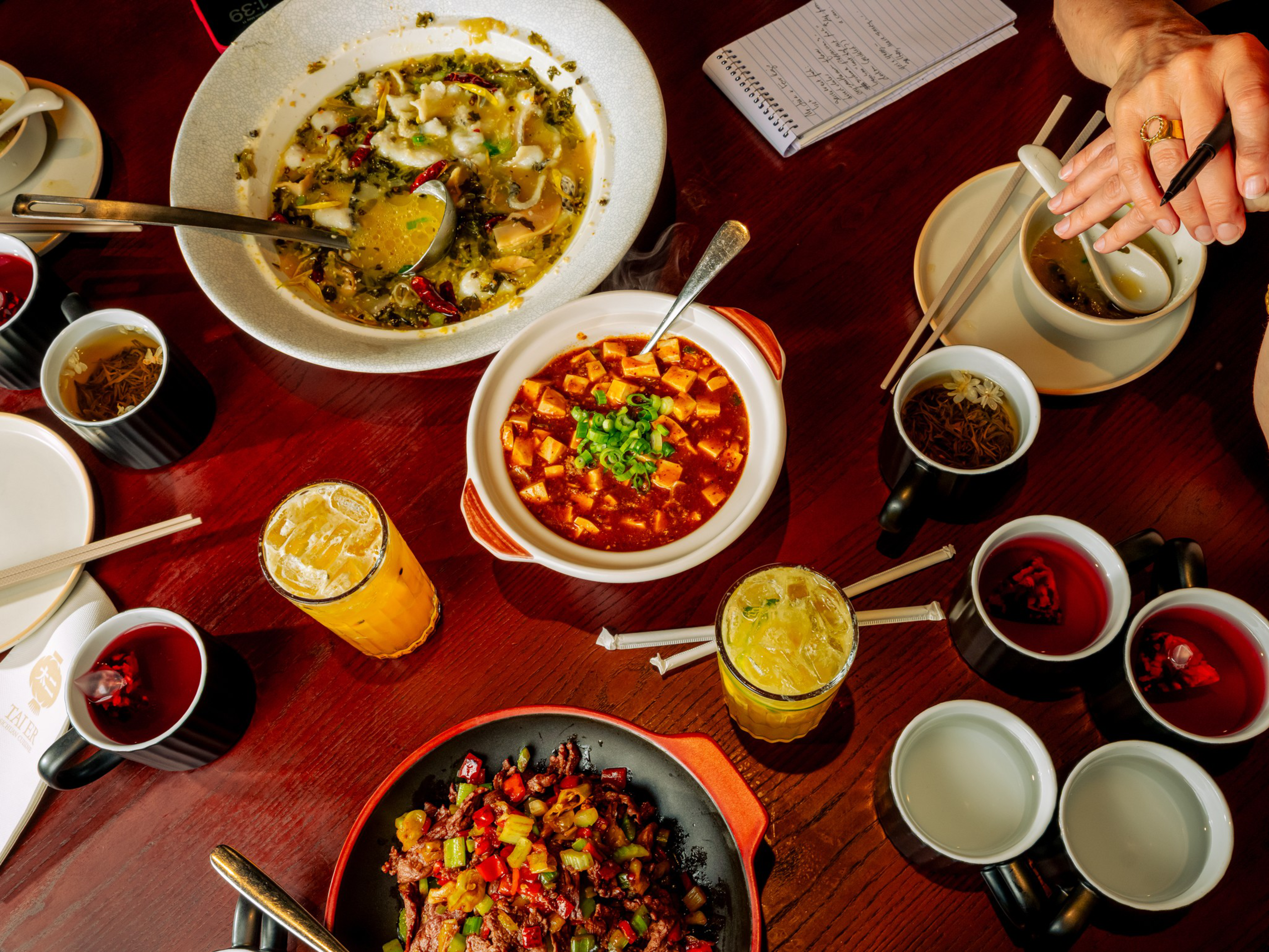 A table with three Chinese dishes, multiple cups of tea, two glasses of iced drinks, chopsticks, a notebook, and a person holding a pen near a cup.
