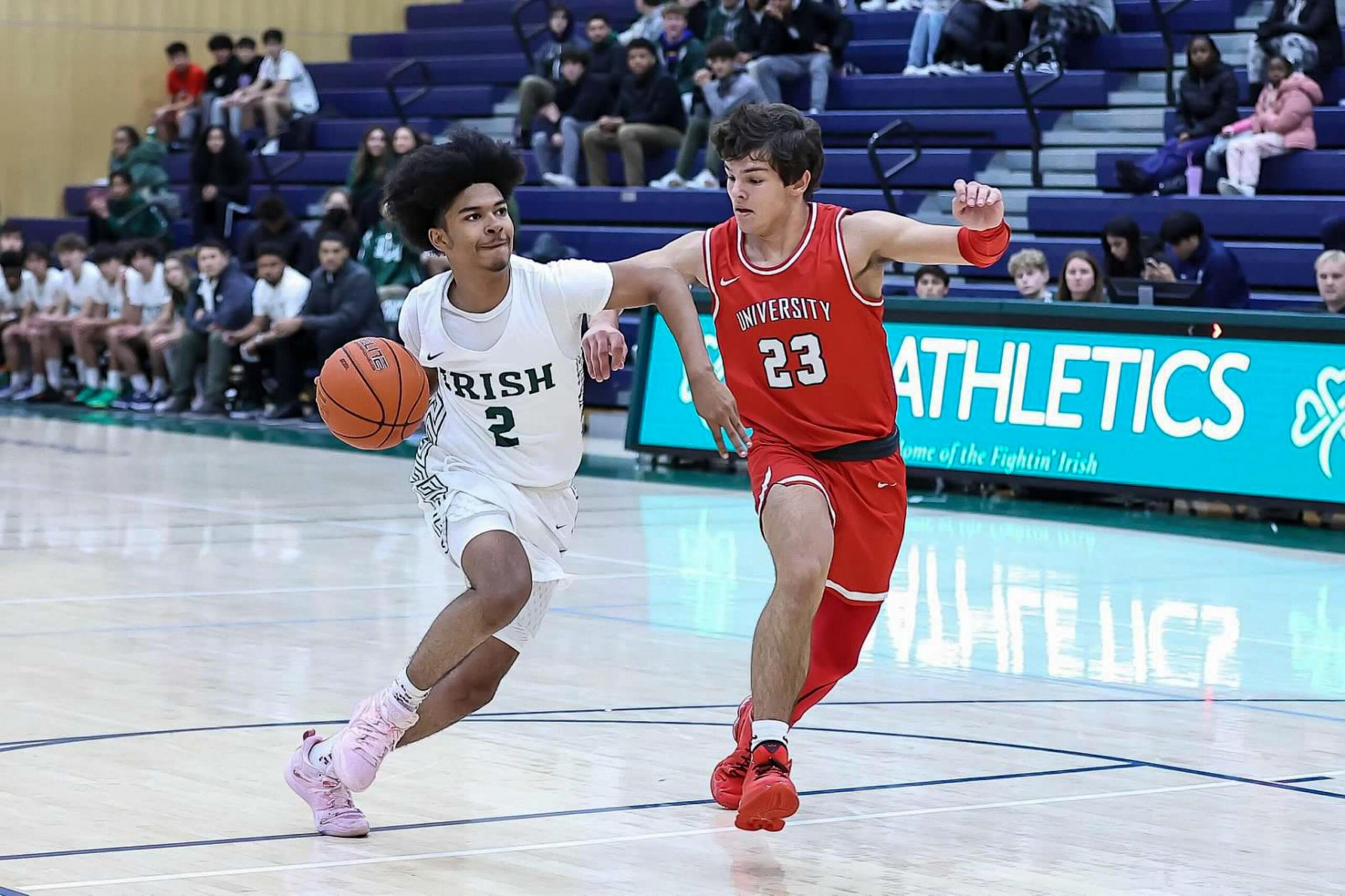 A basketball player in a white "Irish" jersey dribbles while a defender in a red "University" jersey tries to block him on a gym court.