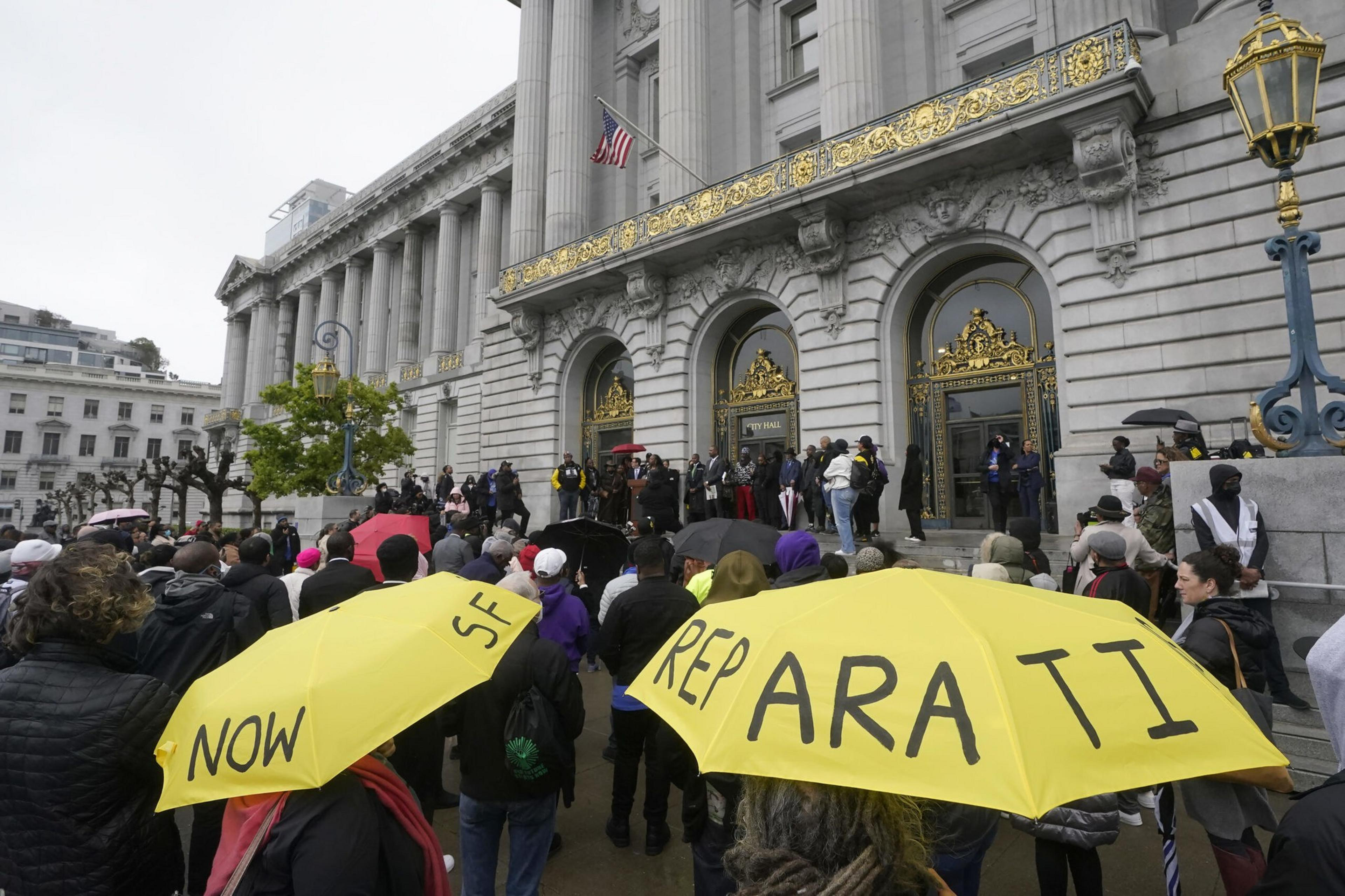 A crowd gathers outside a grand building with gold-trimmed doors, holding yellow umbrellas that read "NOW" and "REPARATI" in black letters.