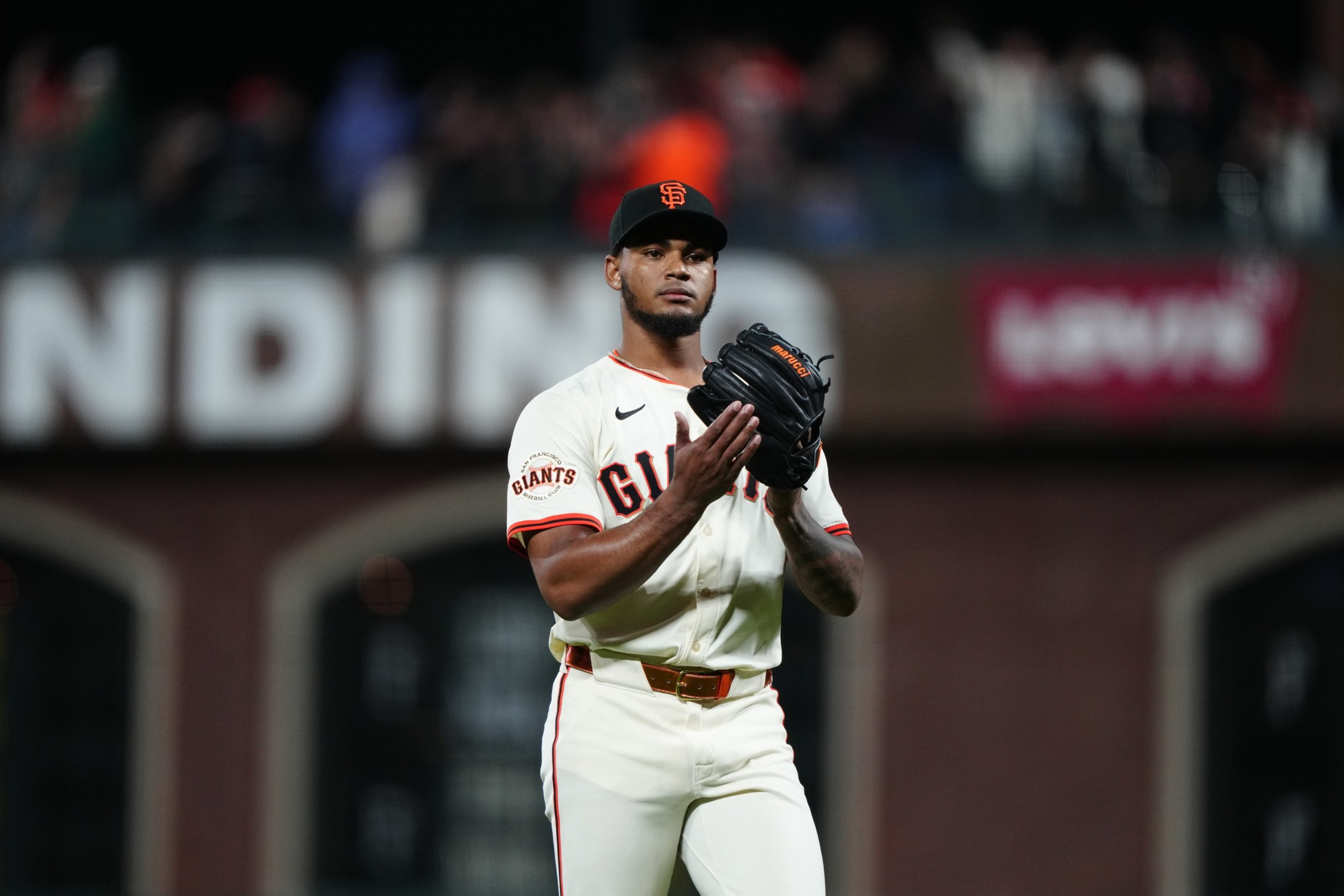 A baseball player wearing a Giants uniform and cap stands on the field, holding a black glove, with blurred spectators and signage in the background.
