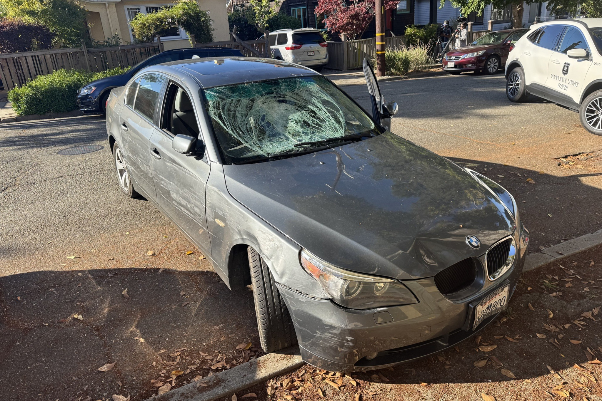 A gray BMW sedan with a severely shattered windshield, dents, and scratches is parked near a curb on a sunny street.