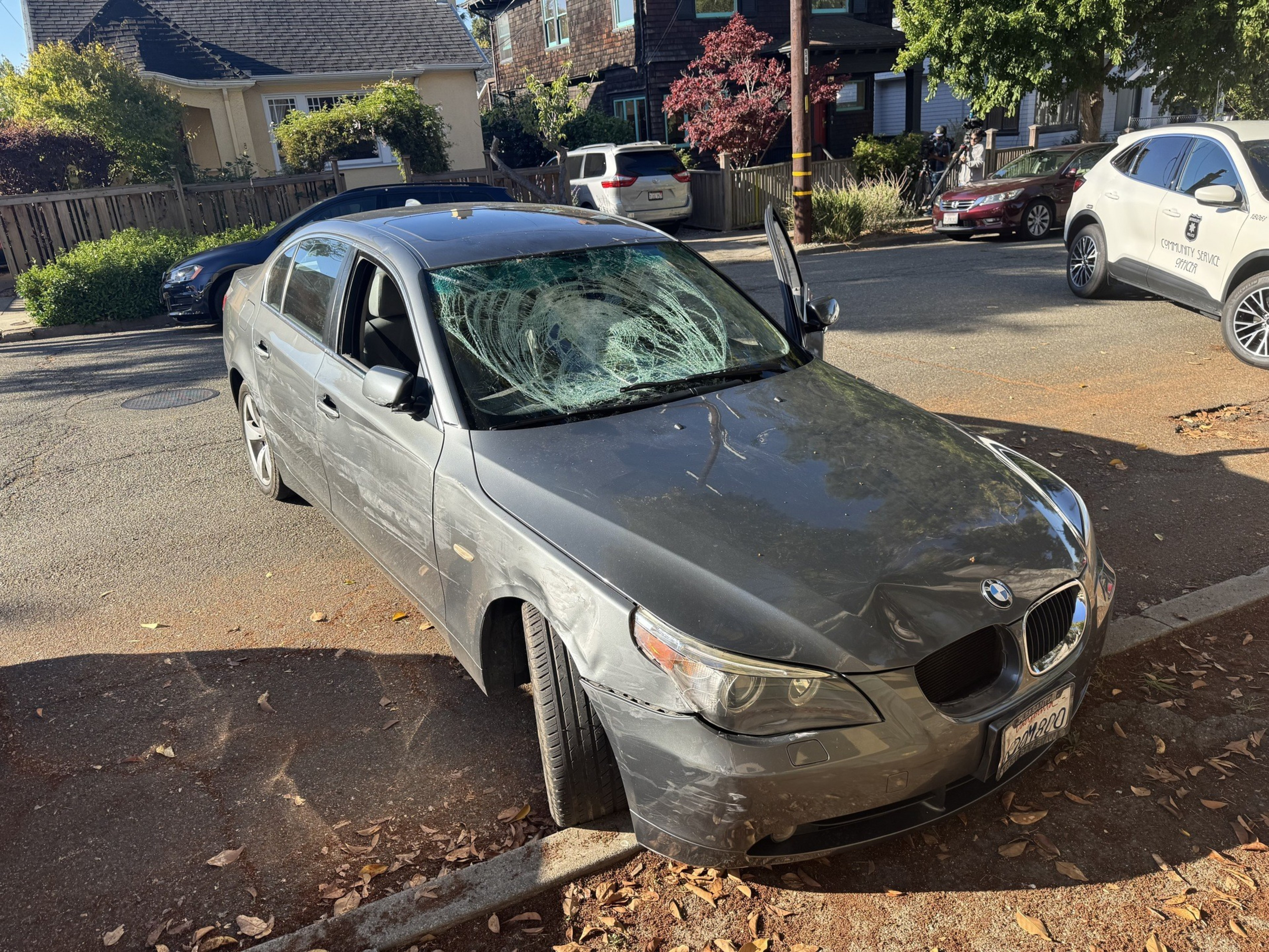 A gray BMW sedan with a severely shattered windshield, dents, and scratches is parked near a curb on a sunny street.