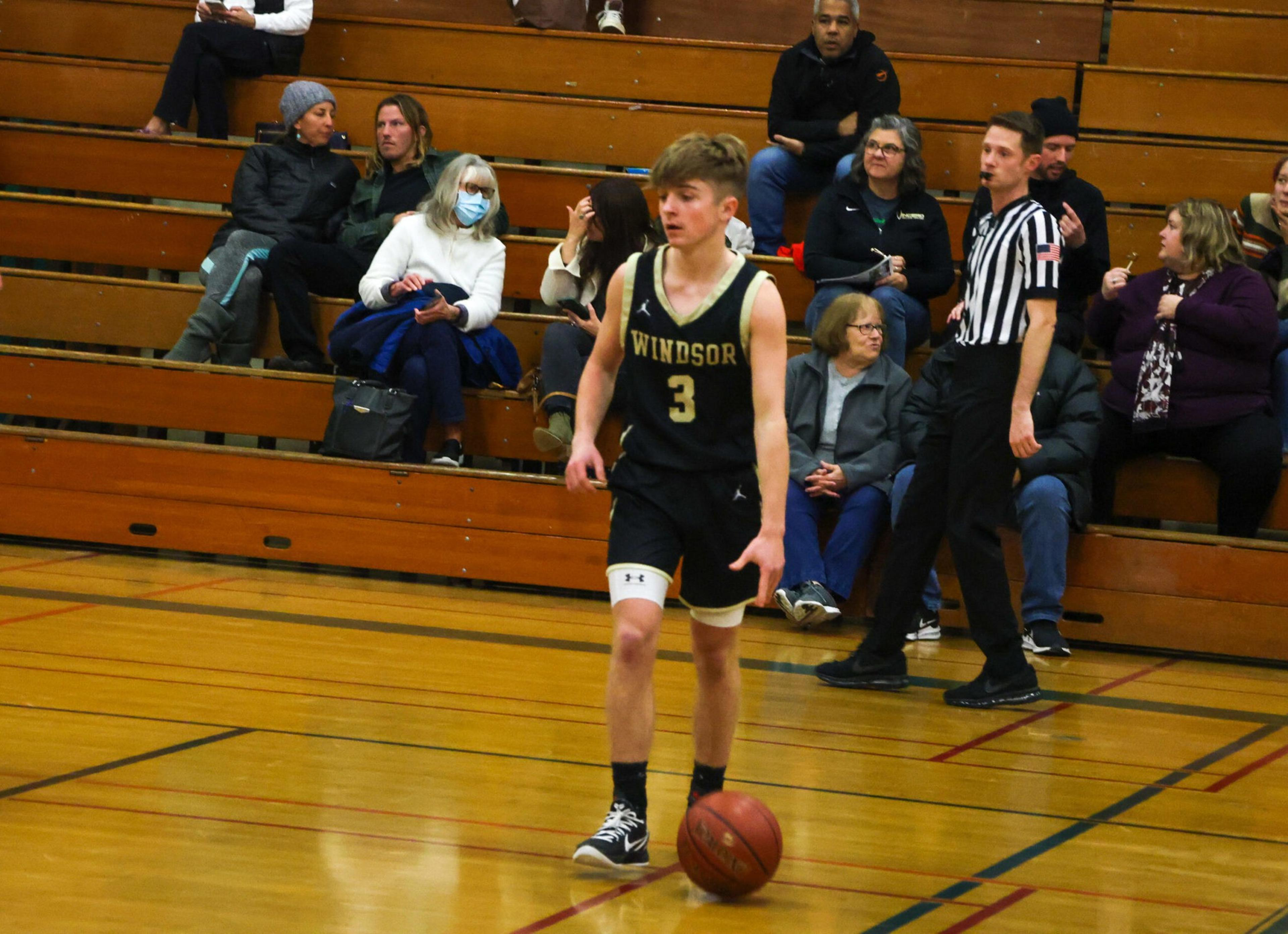 A basketball player in a black Windsor jersey with number 3 dribbles the ball on a gym floor while spectators and a referee watch from wooden bleachers.