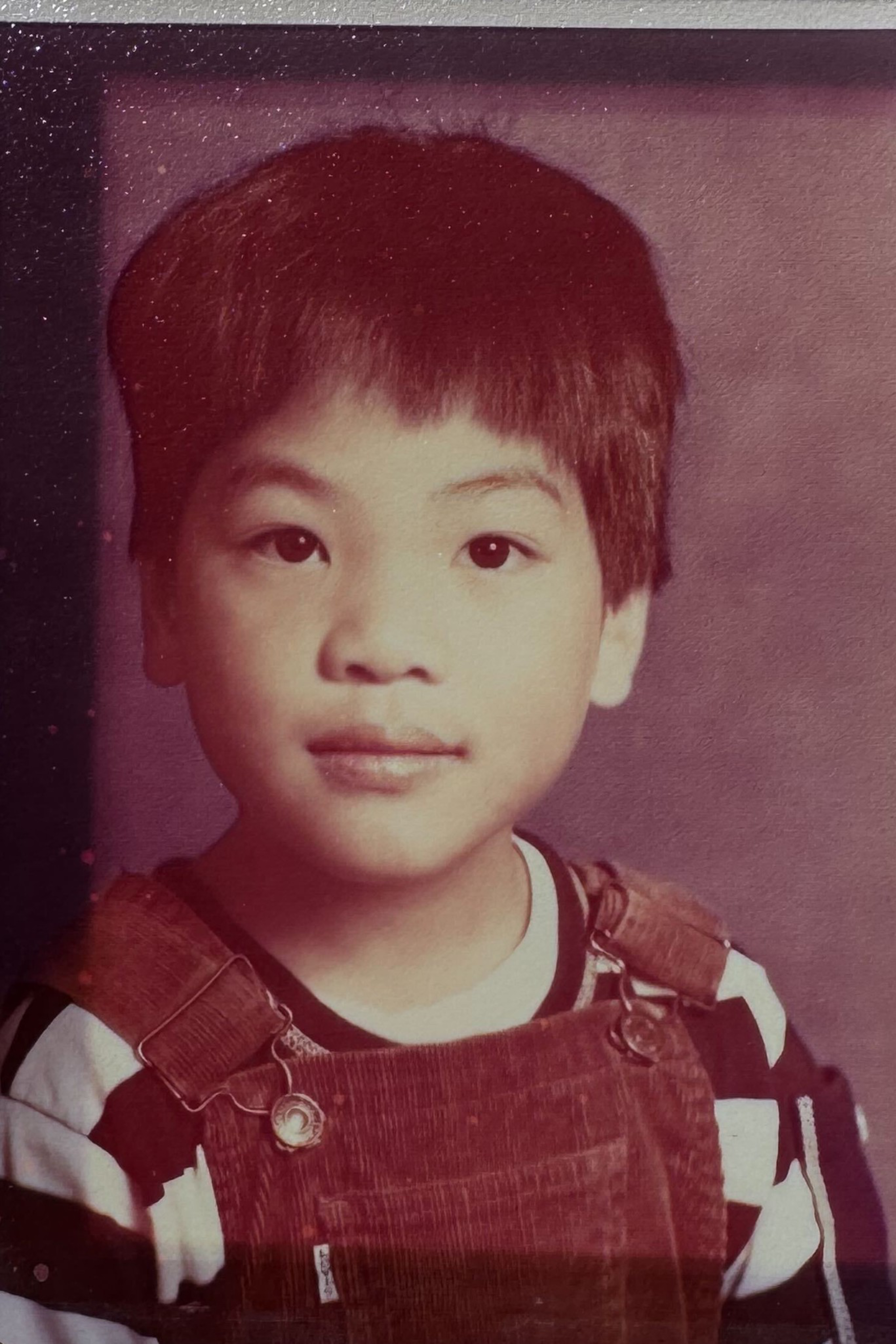 A young child with short, dark hair wears striped shirt and brown corduroy overalls, looking directly at the camera with a calm expression.