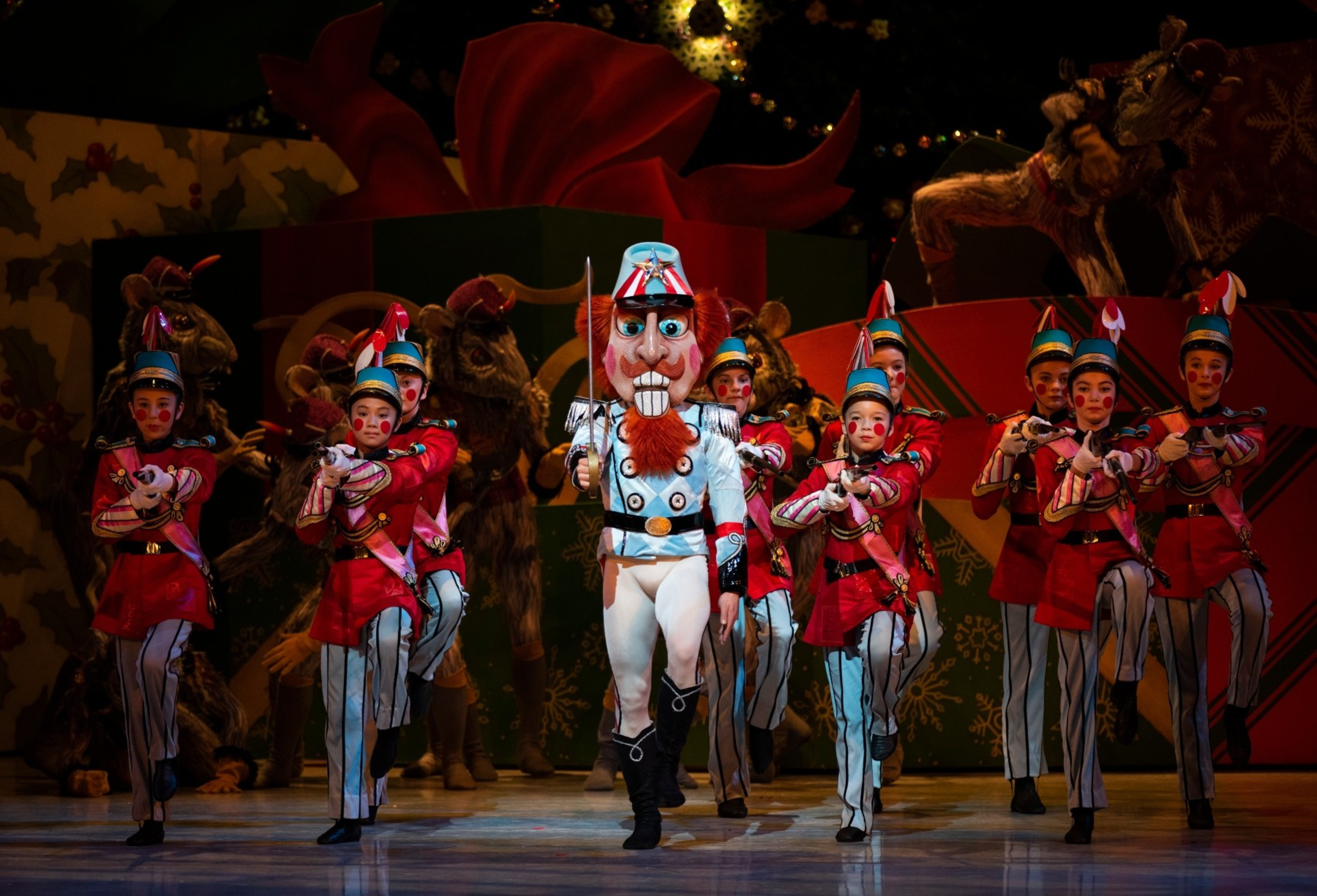 A group of performers dressed as toy soldiers and a large nutcracker figure march on stage, with reindeer and festive decorations behind them.