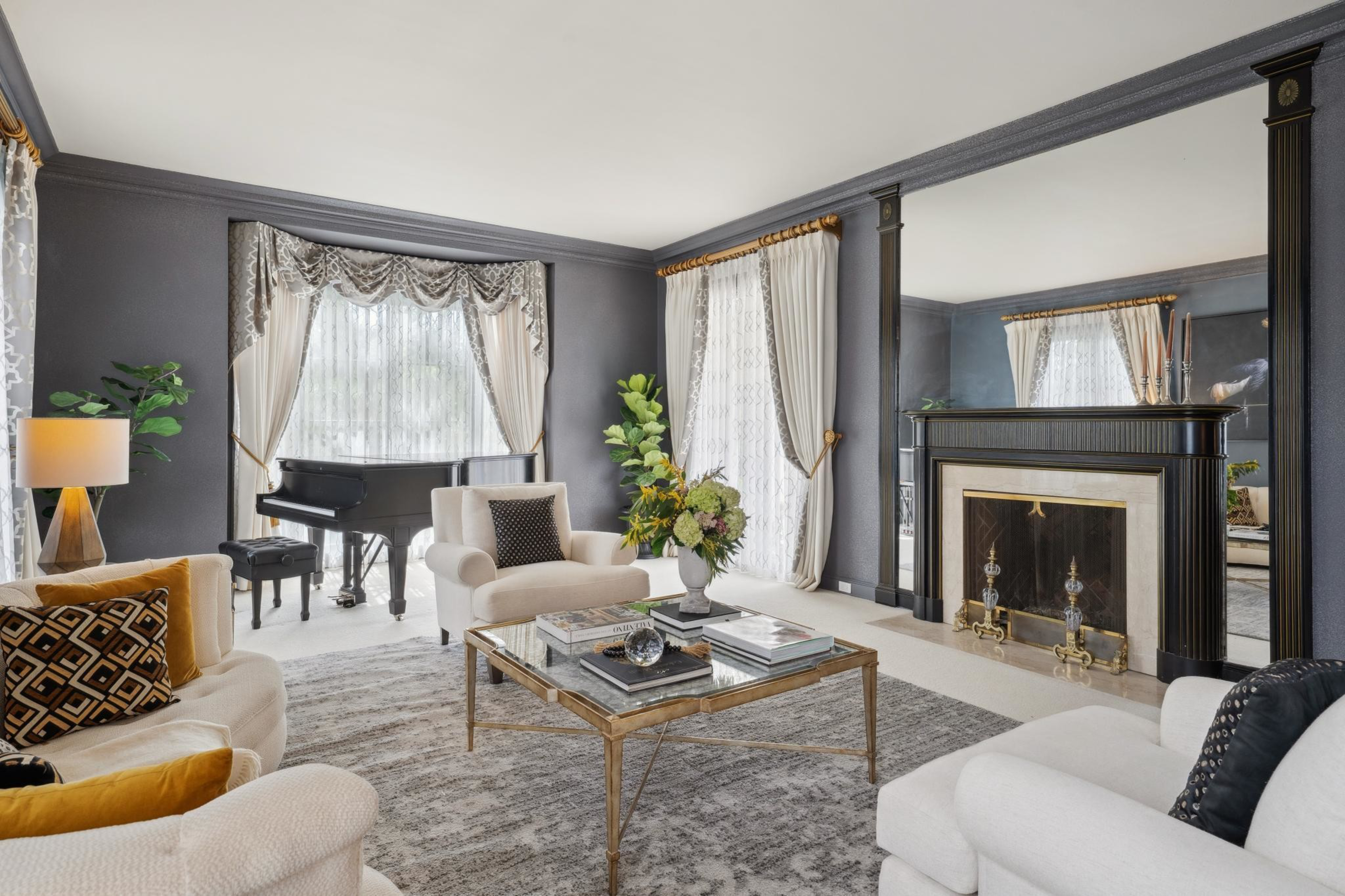 A spacious living room with beige armchairs, a grand piano, a large mirror above a black fireplace, and decorative vases on a glass coffee table.