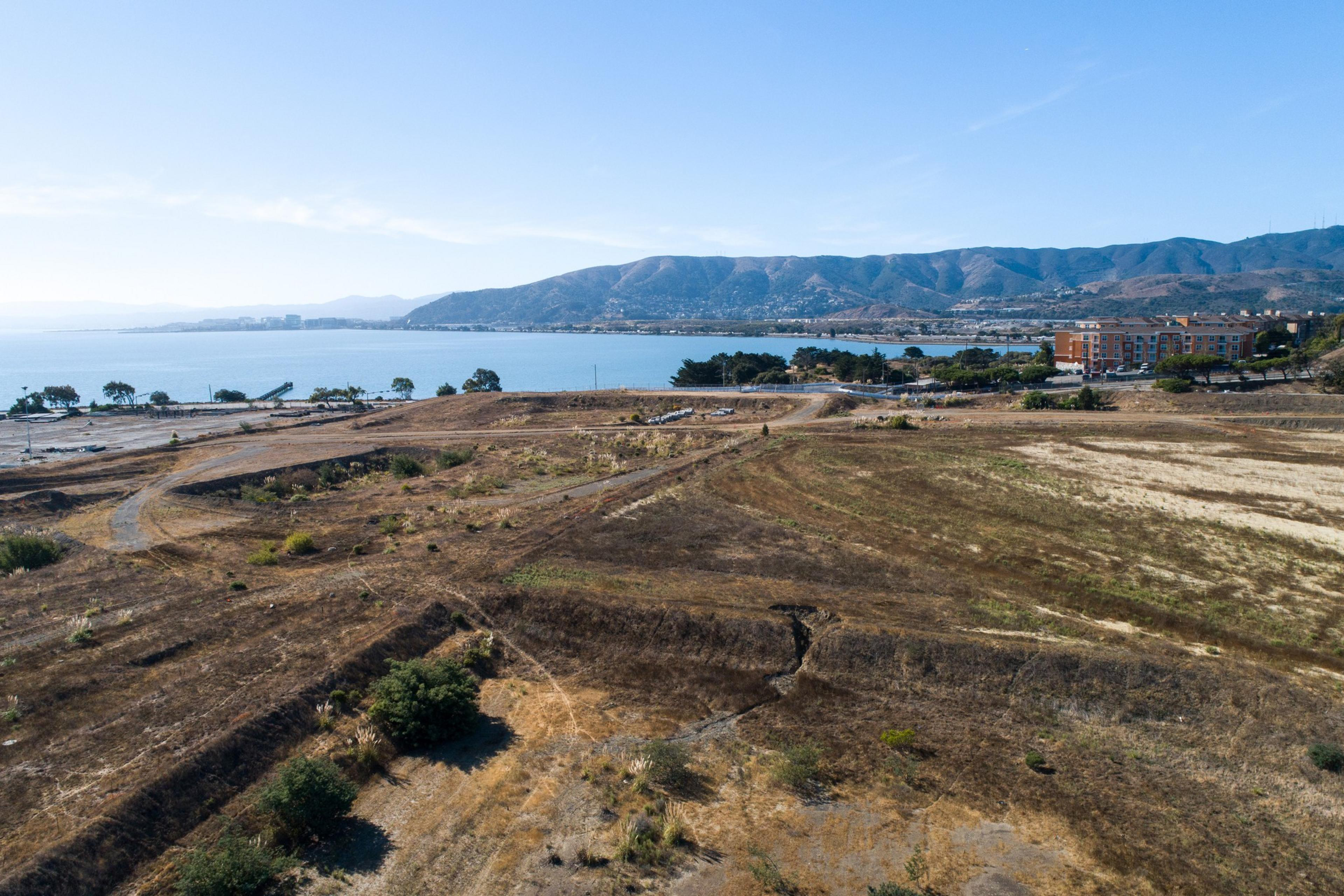 The image shows a coastal landscape with dry, grassy fields in the foreground, a blue bay in the middle, and distant hills under a clear sky.