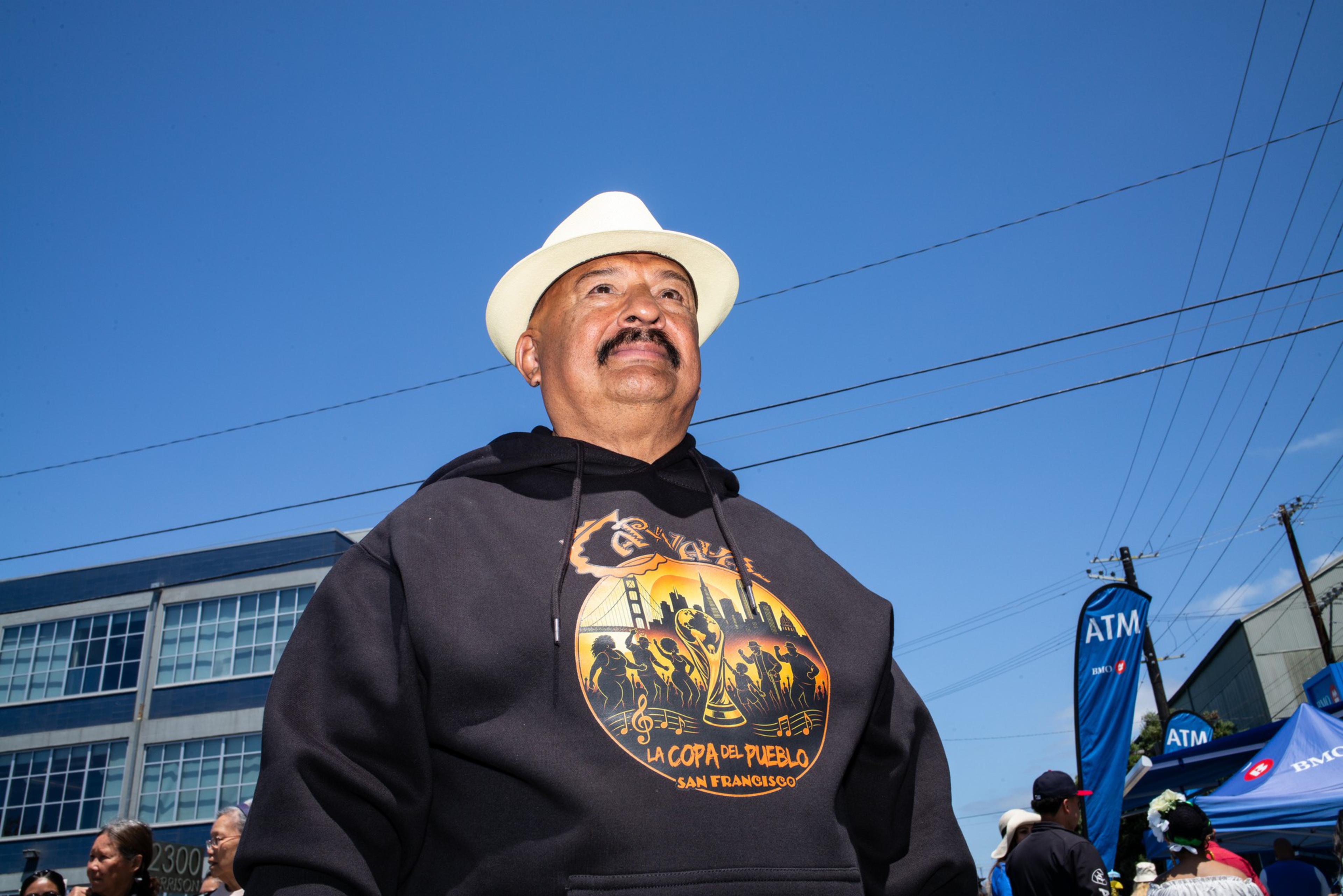 A man wearing a white hat and a black hoodie with a colorful “La Copa de Pueblo San Francisco” design stands outdoors under a clear blue sky.