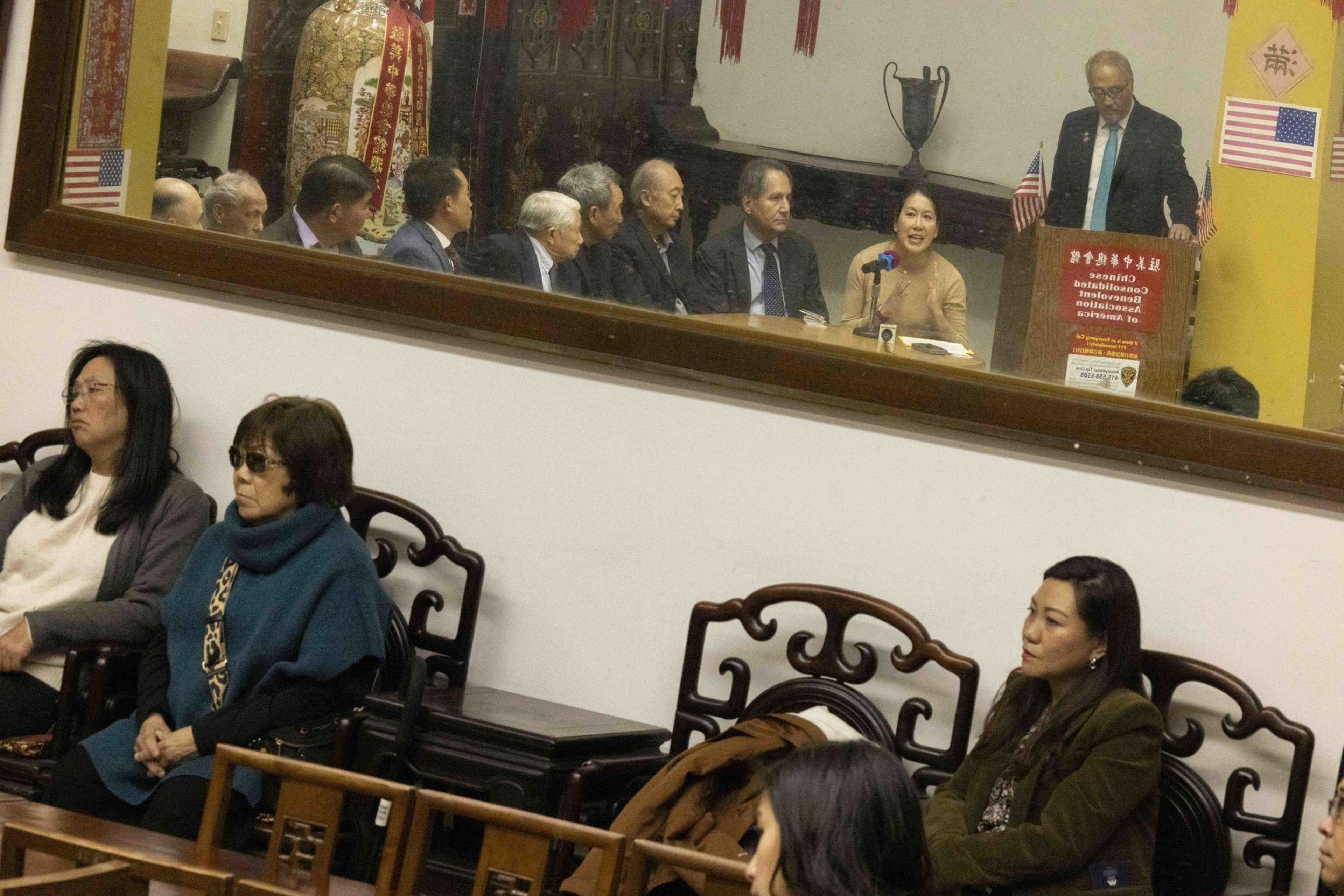 The image depicts people seated in a room with a large mirror reflecting a meeting or discussion behind them. American flags and traditional decor are visible.