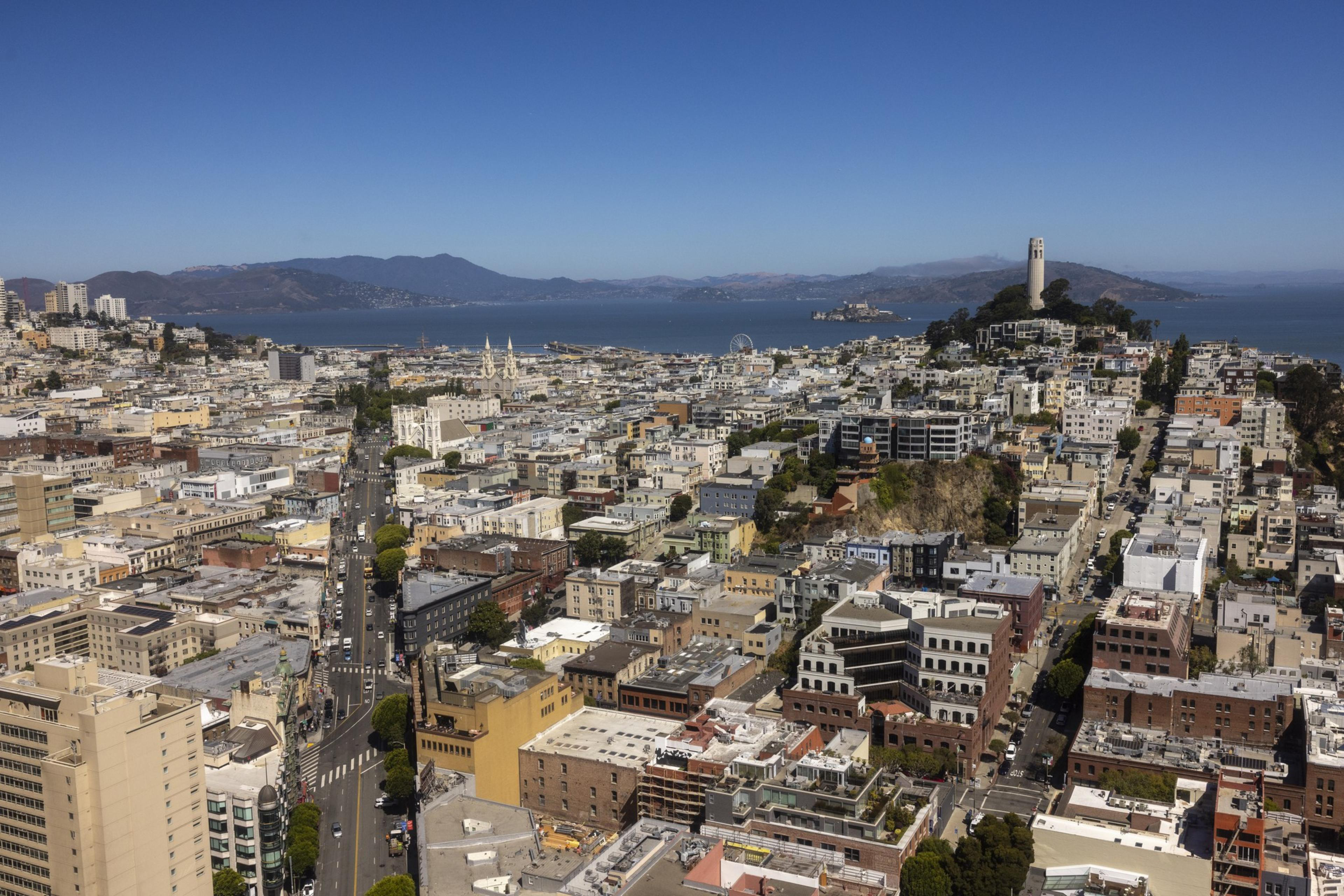 Aerial view of a coastal city with densely packed buildings, a prominent tower on a hill, mountains in the distance, and the ocean with an island and ferris wheel.