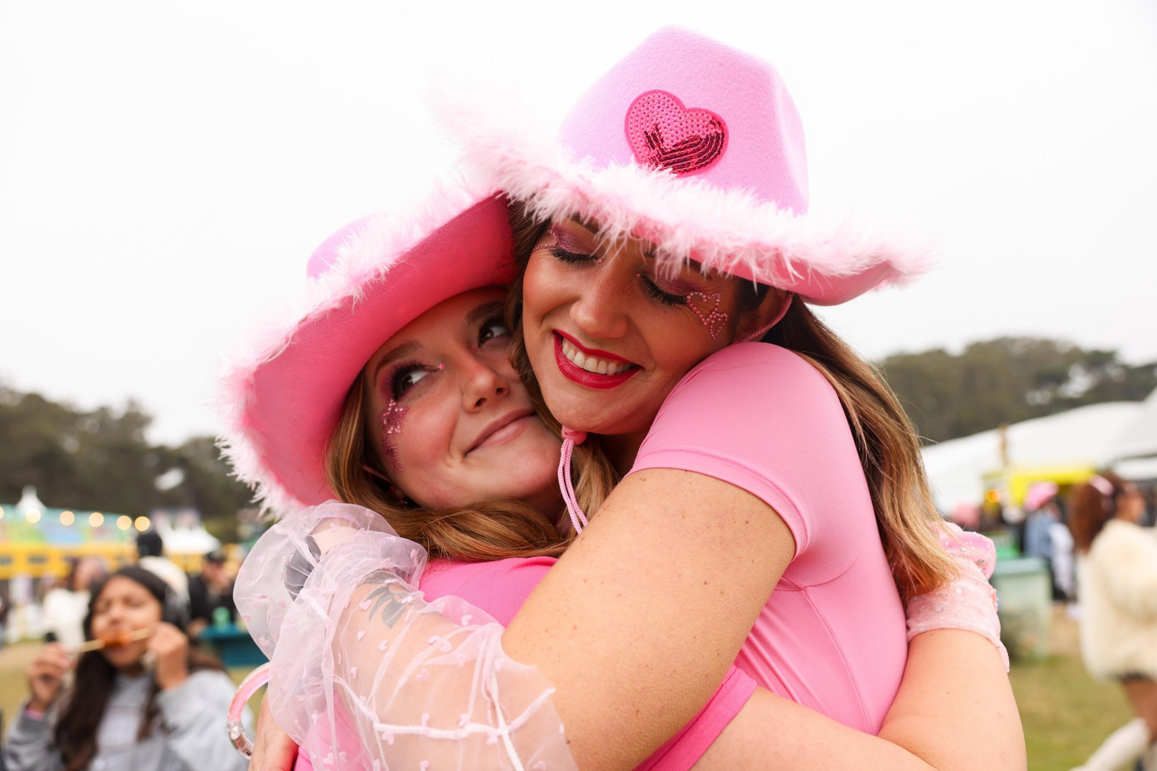 Two people wearing pink outfits and fuzzy pink cowboy hats with heart decorations are hugging and smiling at an outdoor event.