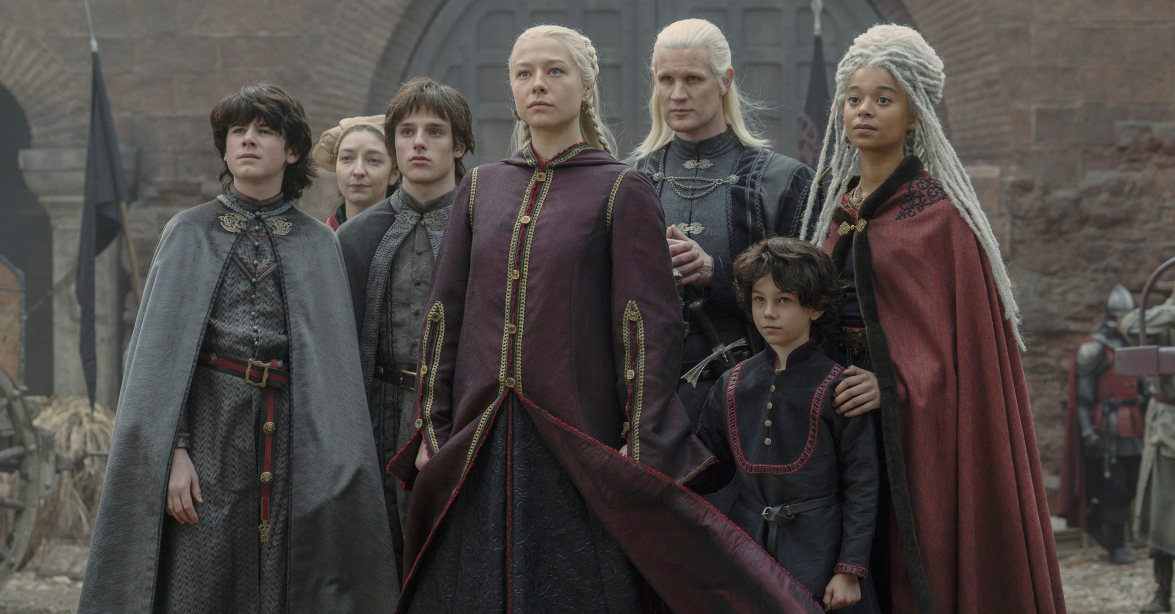 Seven people dressed in medieval fantasy clothing stand solemnly in a courtyard with stone walls and an arched doorway behind them.
