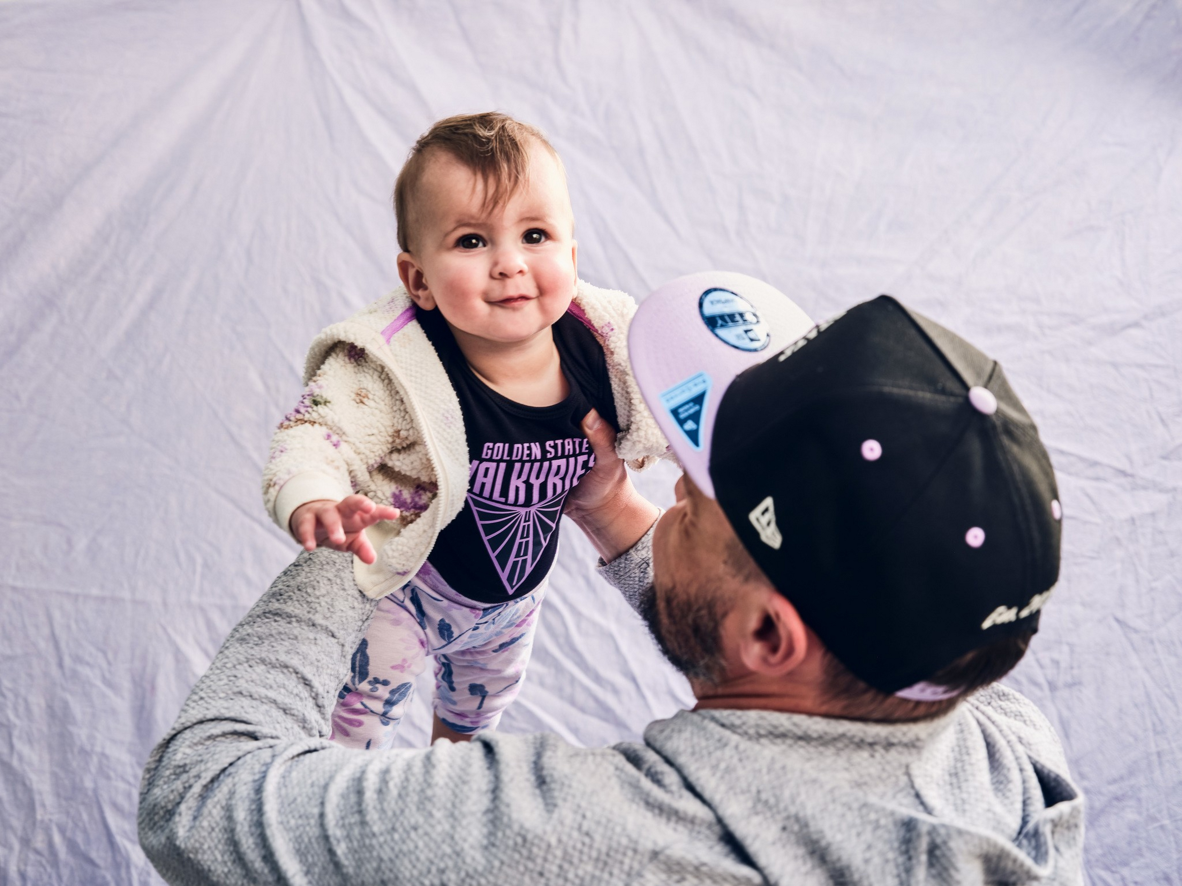 A man wearing a black and white cap lifts a smiling baby dressed in a purple shirt and floral pants against a plain light background.
