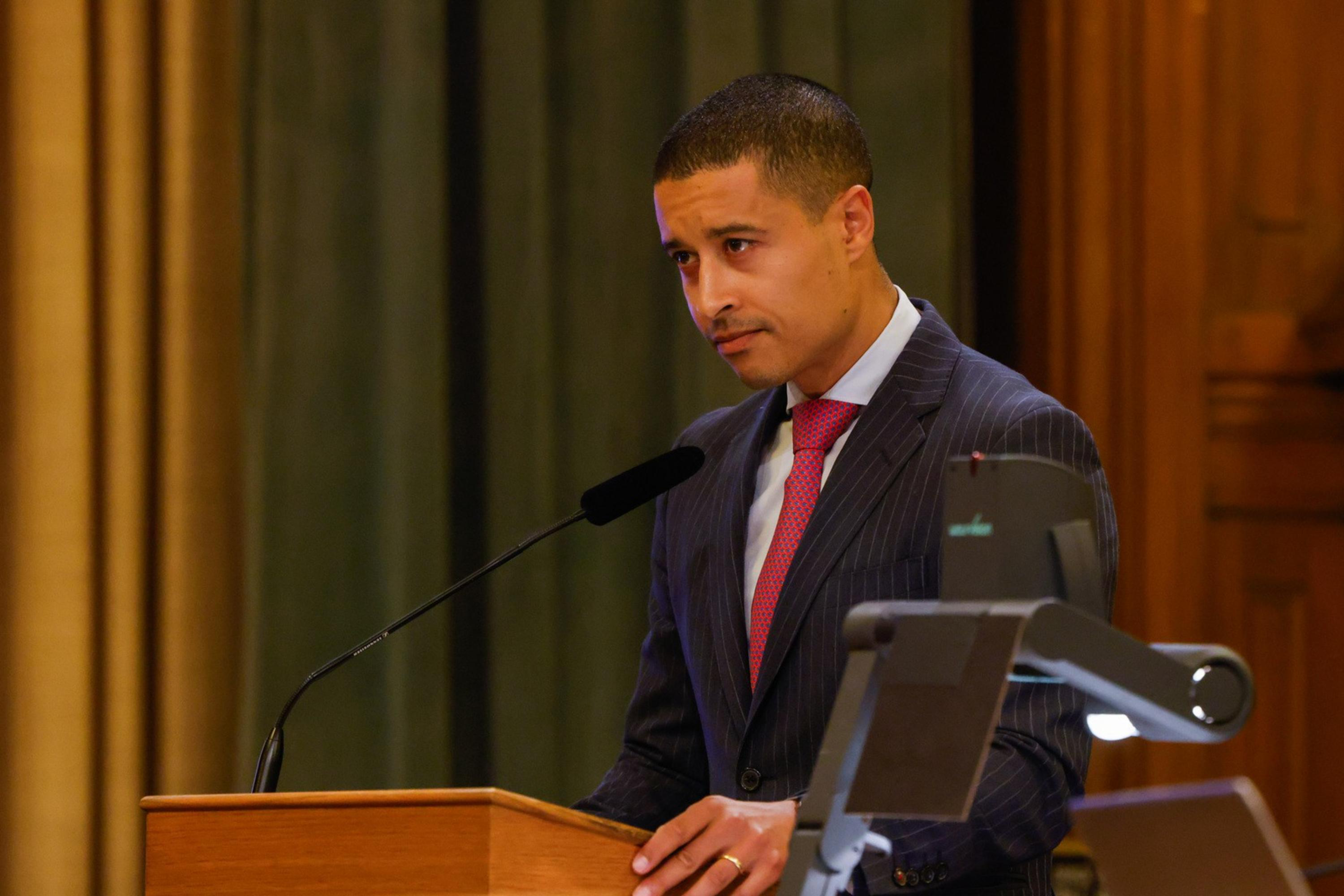 A person in a suit speaks at a wooden podium with a microphone. The setting appears formal, likely a conference or meeting room, with a laptop nearby.