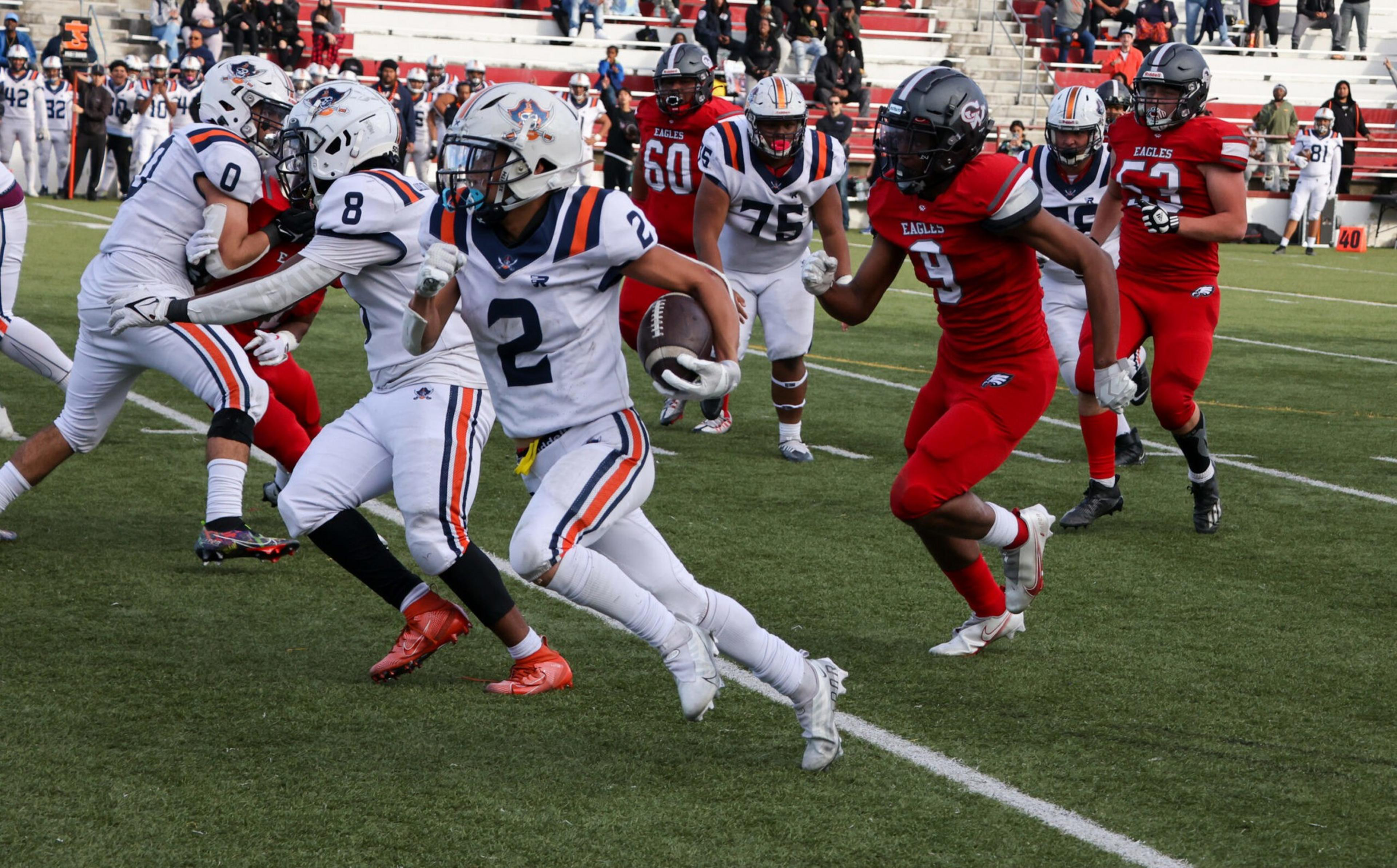 A football player in white with number 2 runs with the ball while players in red attempt to catch him on a green field.