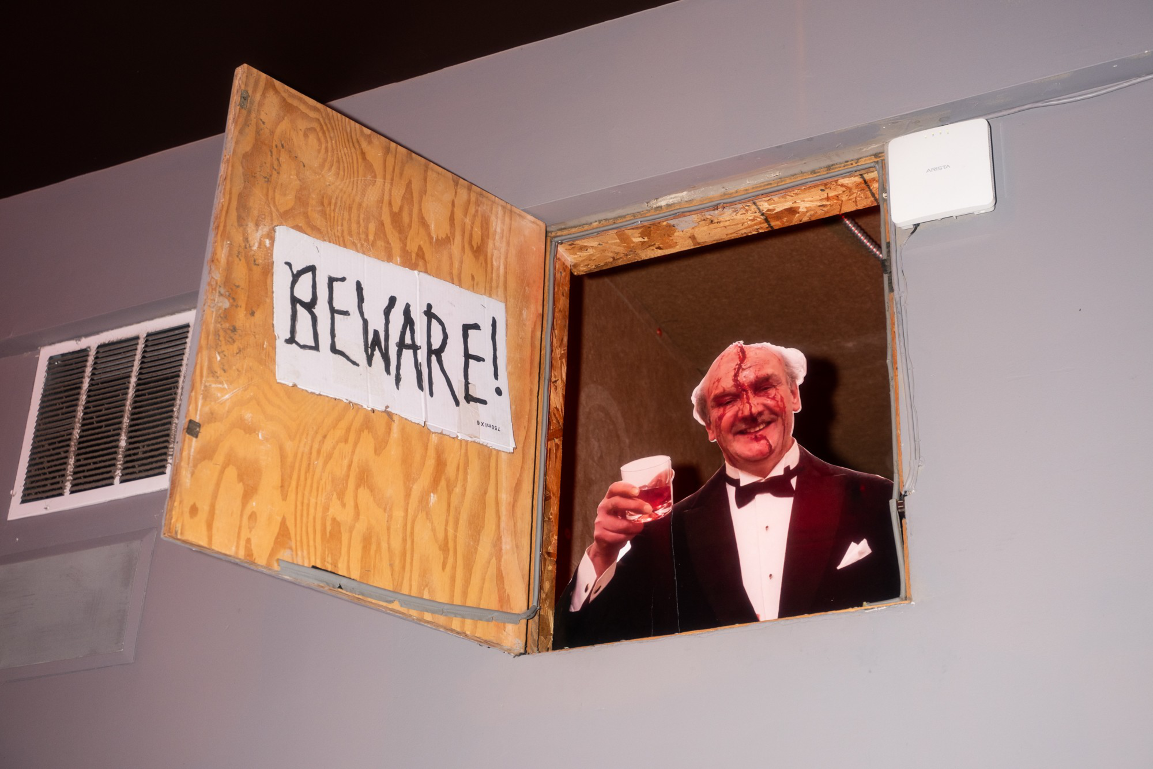A wooden hatch door with a "BEWARE!" sign opens to reveal a man in a tuxedo with a bloody face holding a glass of red liquid.