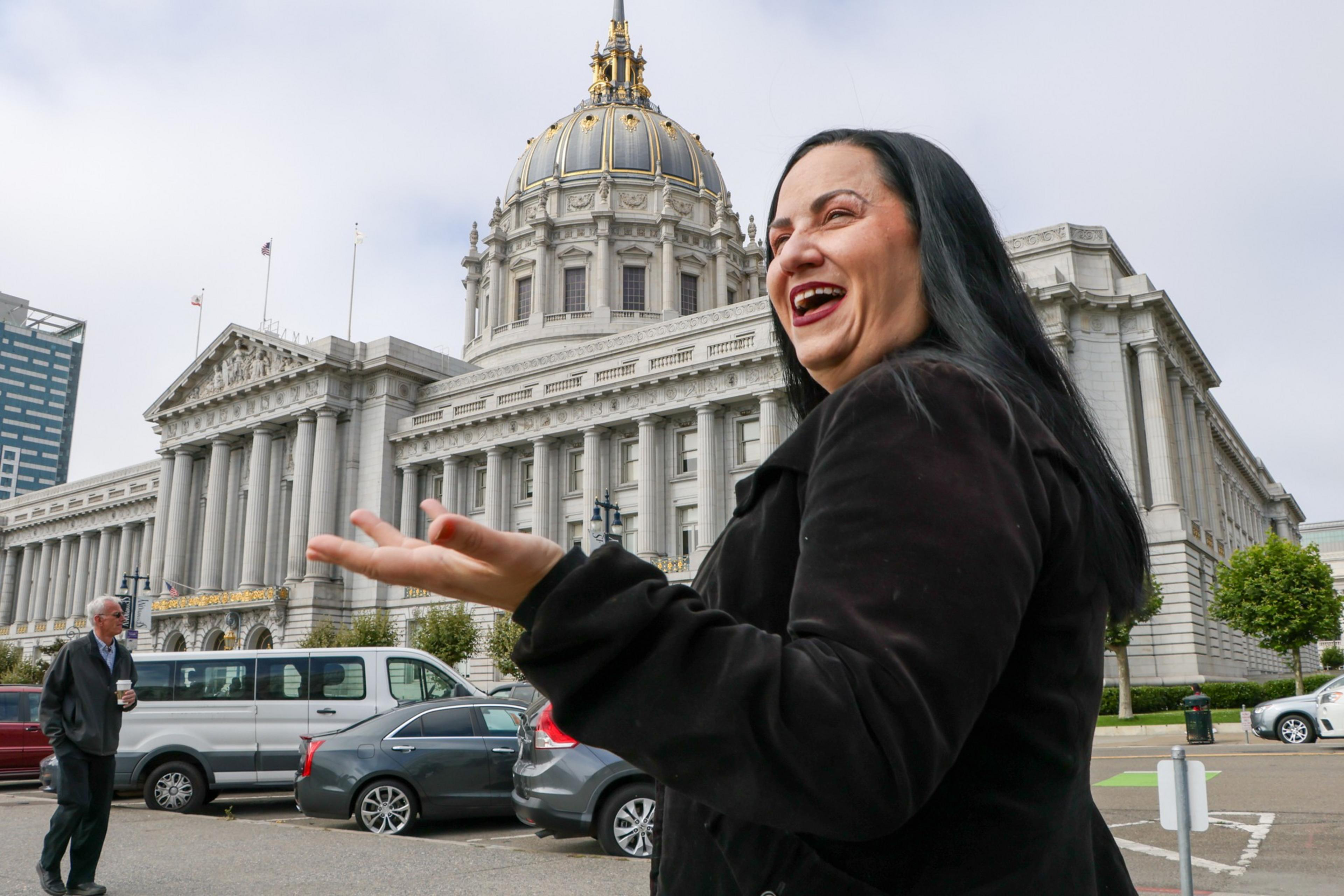 A woman with long black hair wearing a black coat laughs joyfully with a grand neoclassical building featuring a gold-domed roof in the background.