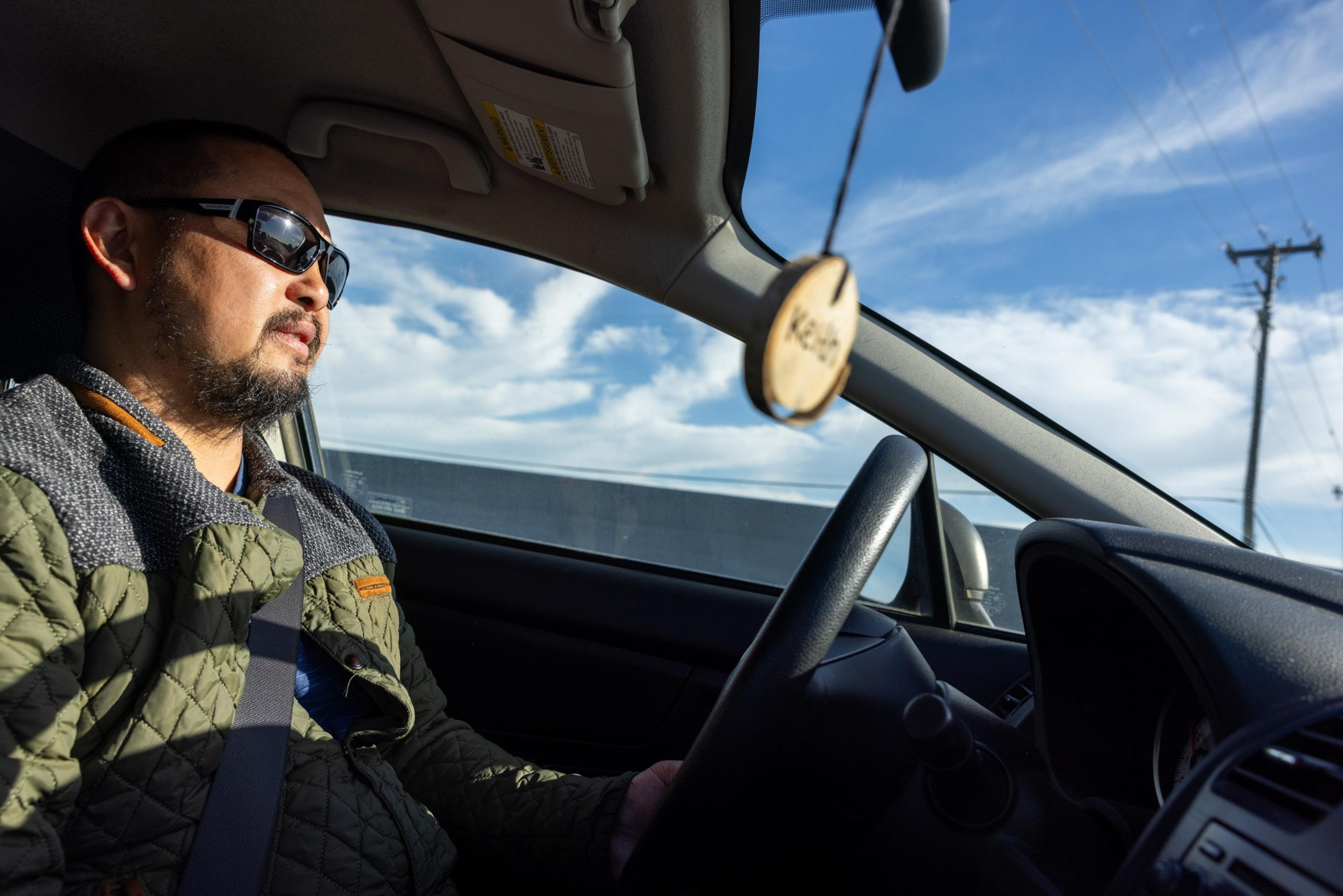 A man wearing sunglasses and a green quilted jacket drives a car. A wooden pendant with &quot;Kalos&quot; hangs from the rearview mirror. The sky is clear and blue.