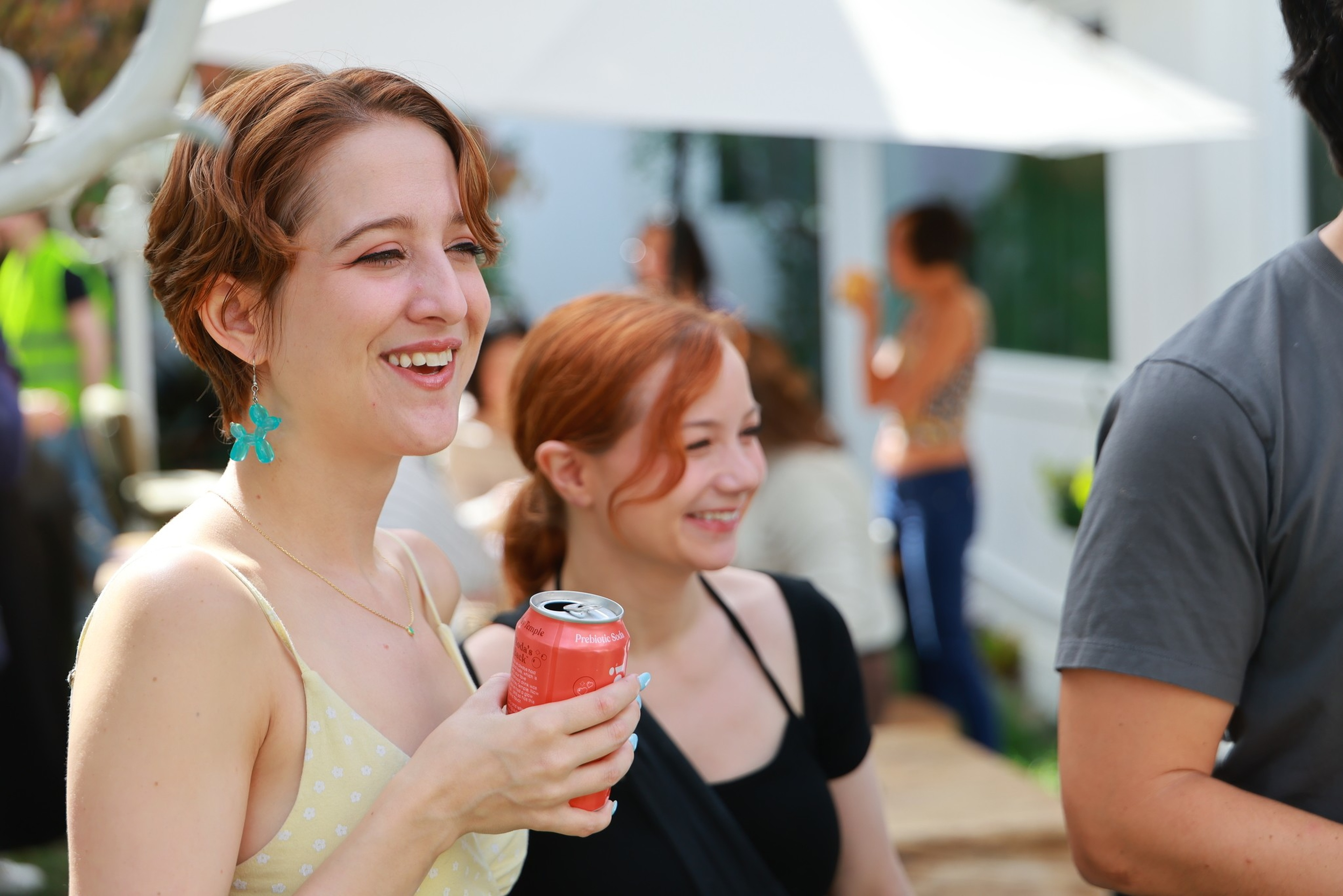 Two young women are smiling and enjoying a social outdoor gathering, with one holding a red canned drink and people mingling in the background.
