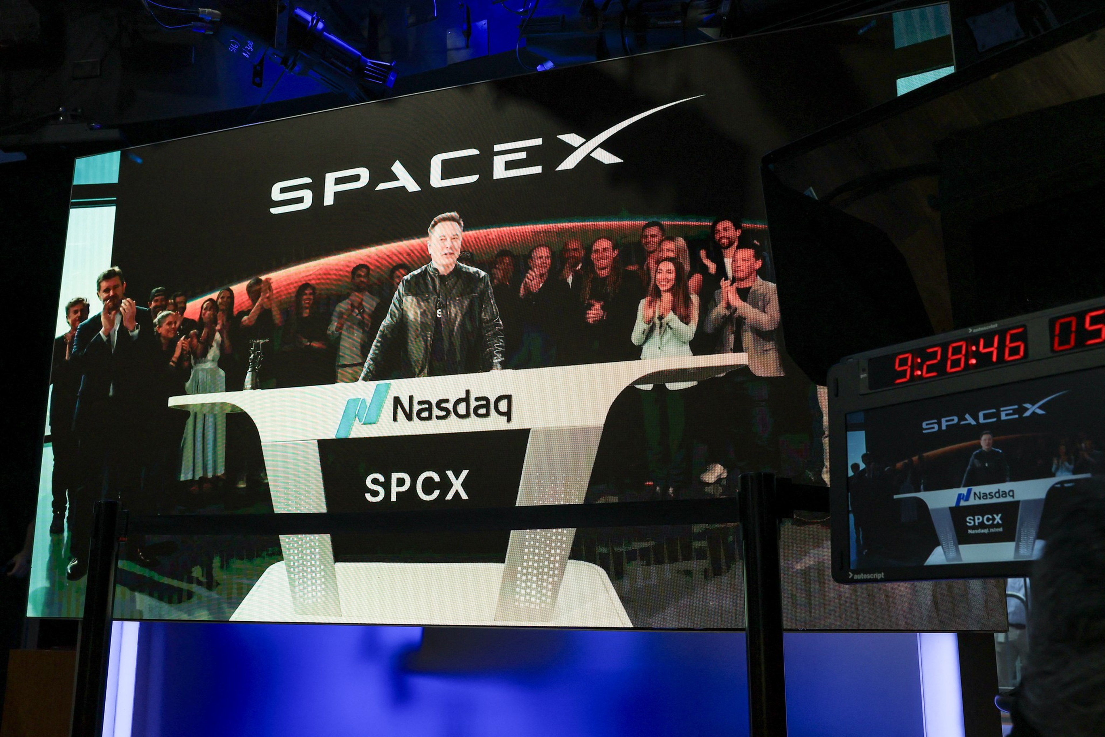 Elon Musk stands at a Nasdaq podium labeled SPCX, with a large SpaceX logo behind him and people clapping in the background.