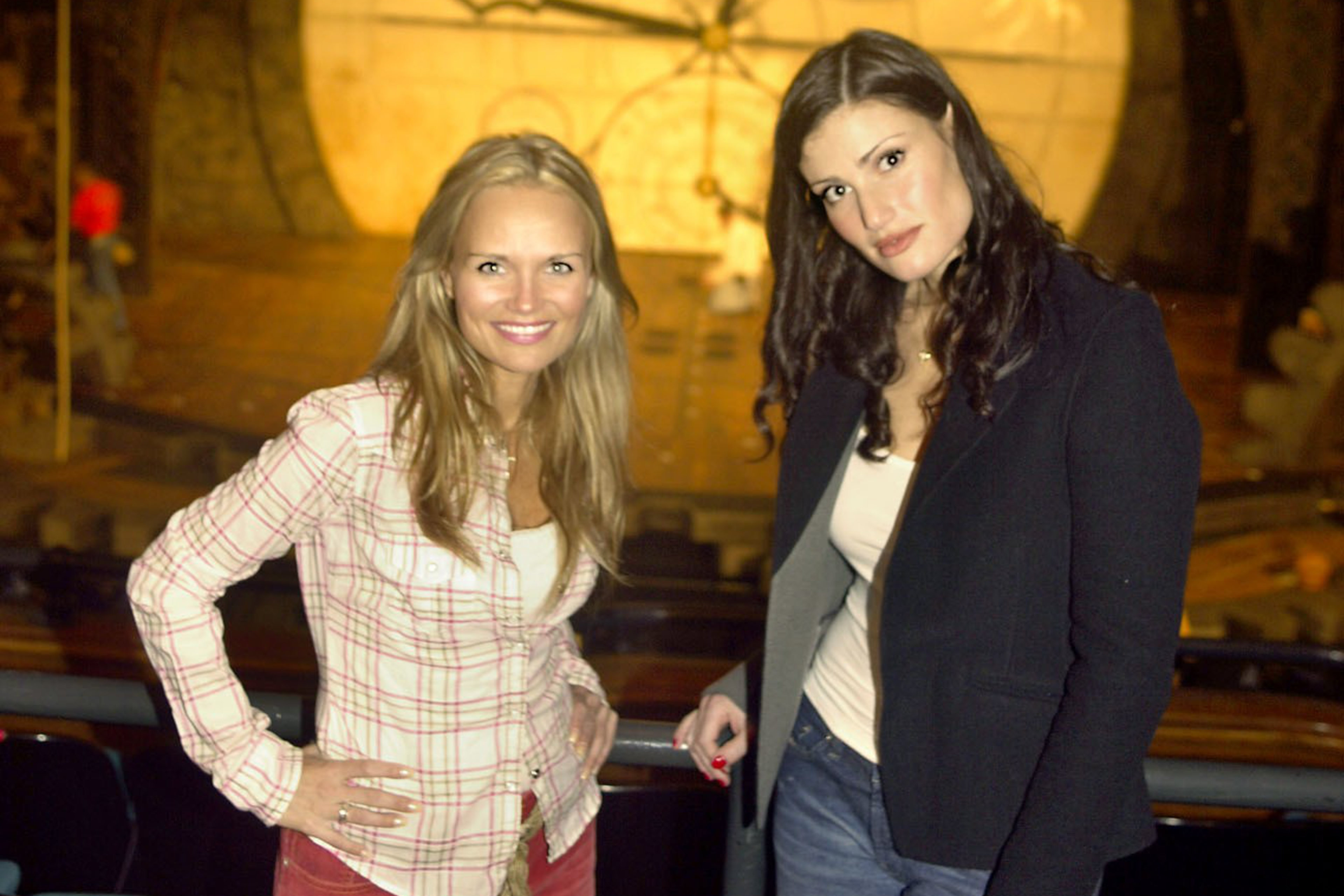 Two women stand smiling in front of a theater stage with a large clock and dragon decoration above. The ambience suggests a set for a theatrical production.