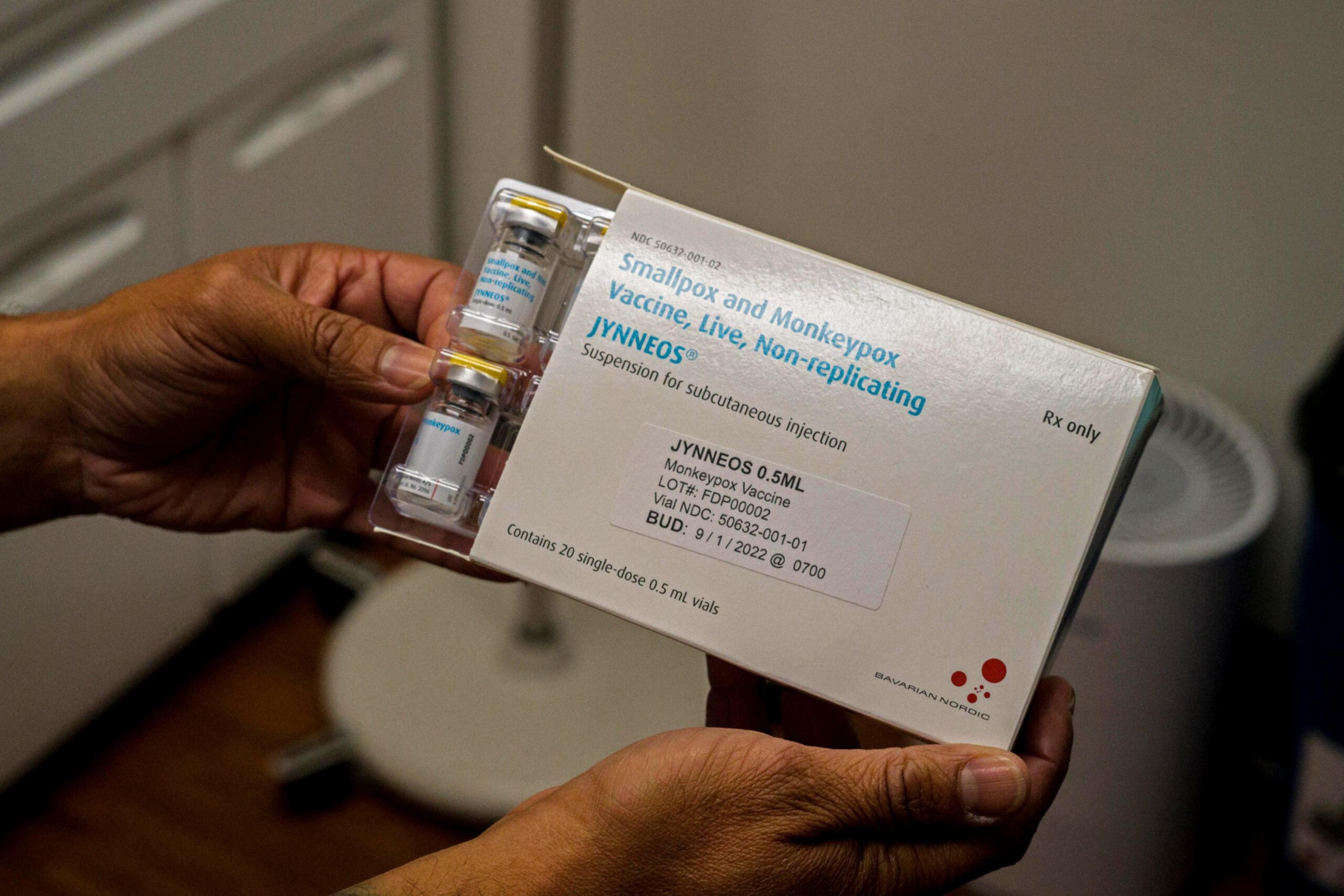 Hands hold a box labeled "Smallpox and Monkeypox vaccine, Live, Non-replicating JYNNEOS" containing two vials of the vaccine for subcutaneous injection.