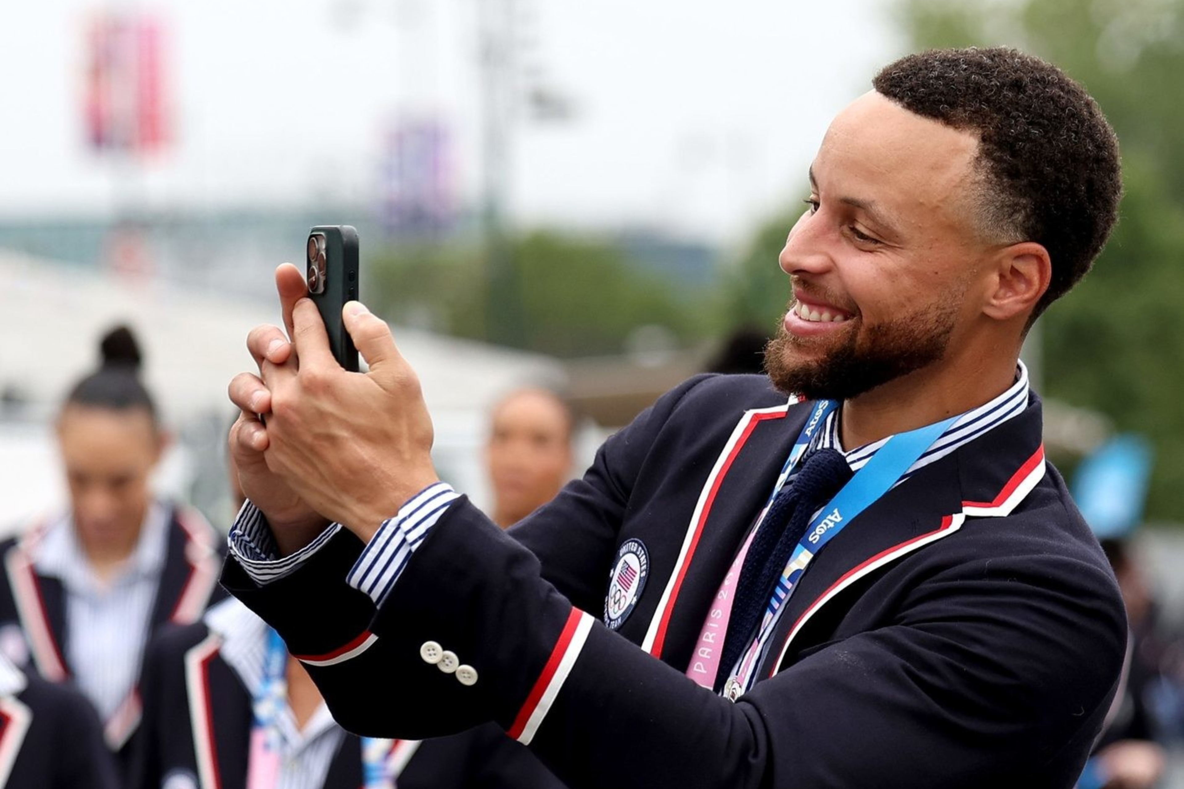 A group of people in matching navy blazers with red and white trim, some wearing blue jeans, are walking outdoors; one man is taking a selfie.