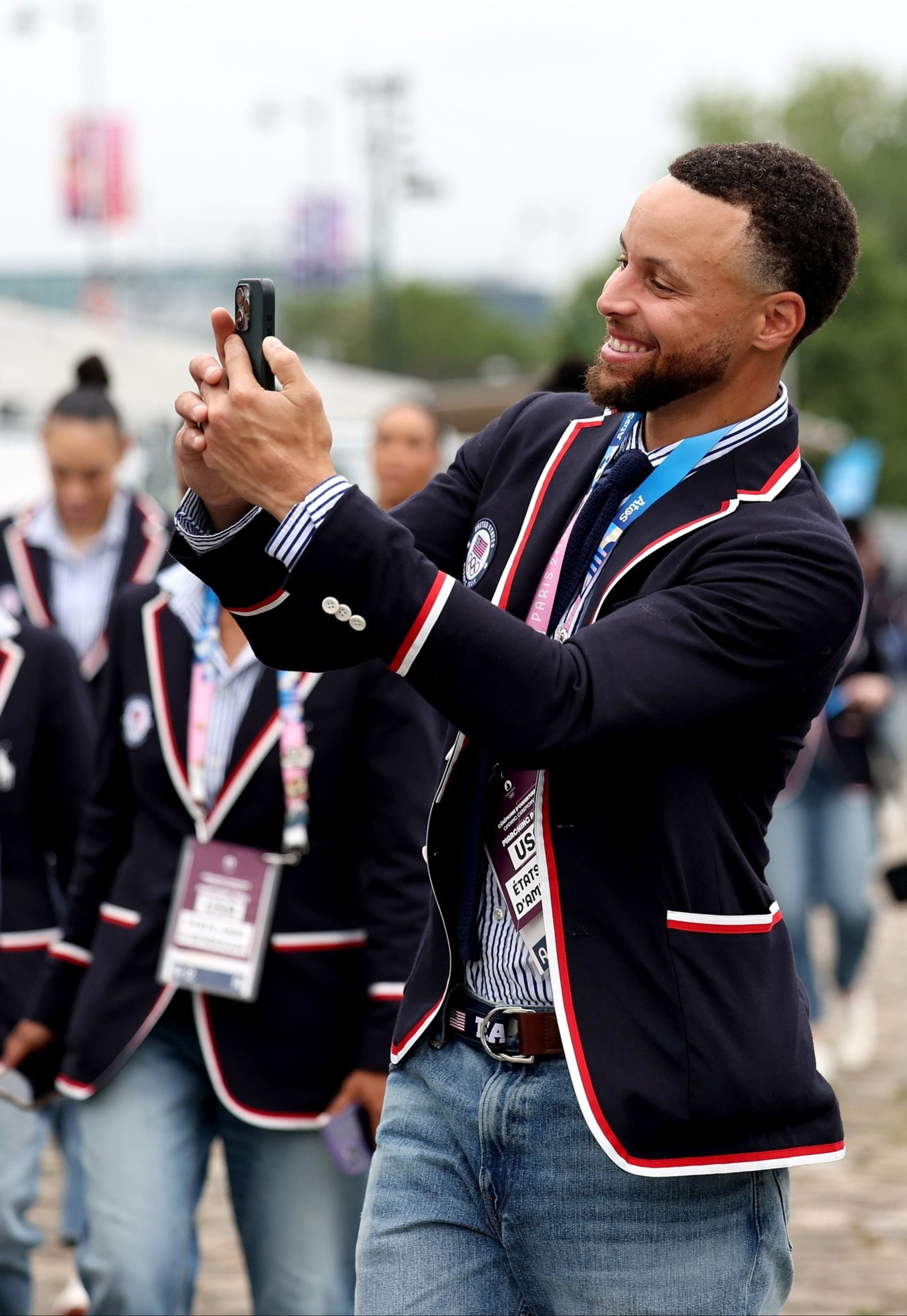 A group of people in matching navy blazers with red and white trim, some wearing blue jeans, are walking outdoors; one man is taking a selfie.