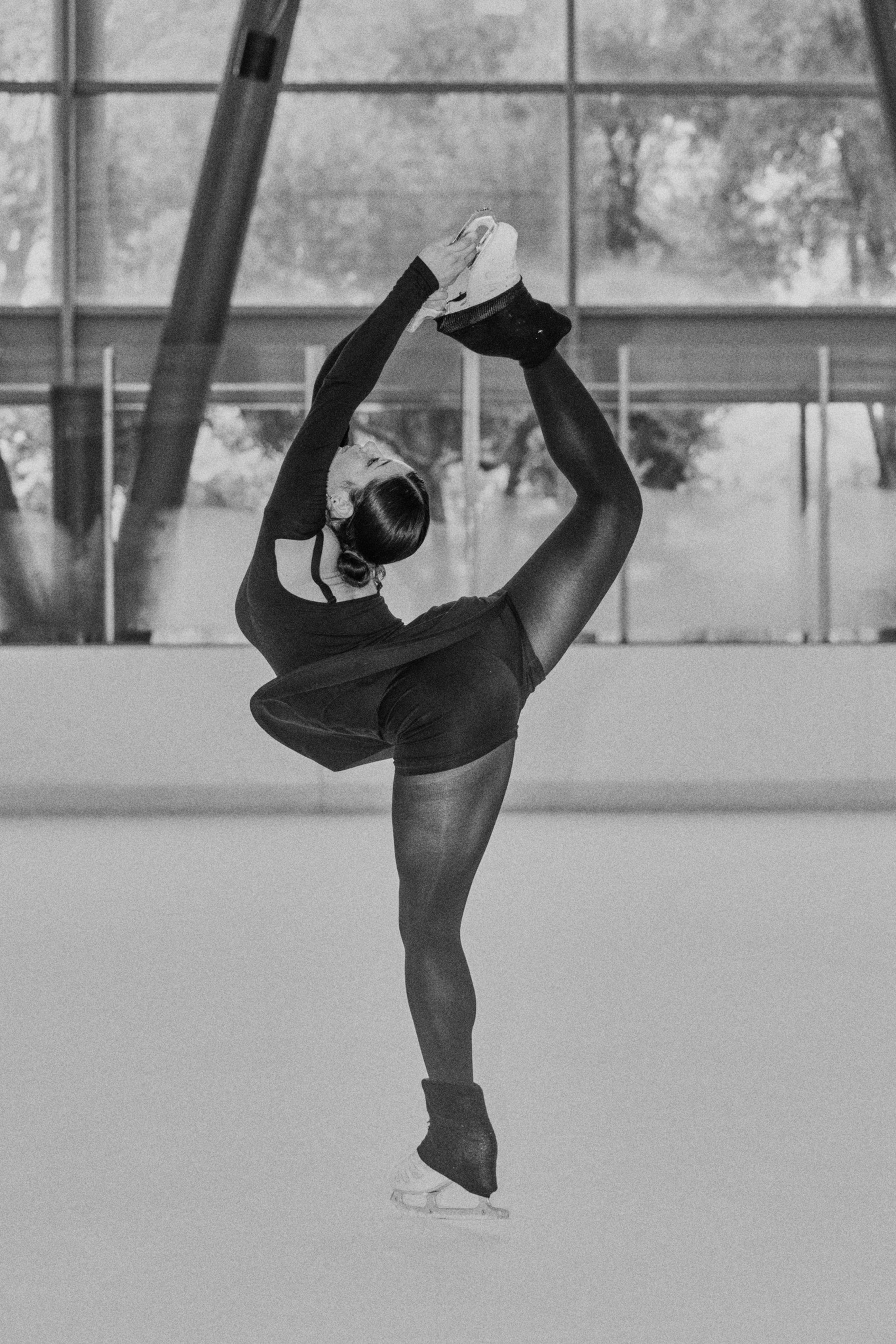 A figure skater in a black outfit balances on one skate while holding her raised leg behind her head in a deep backbend on the ice rink.
