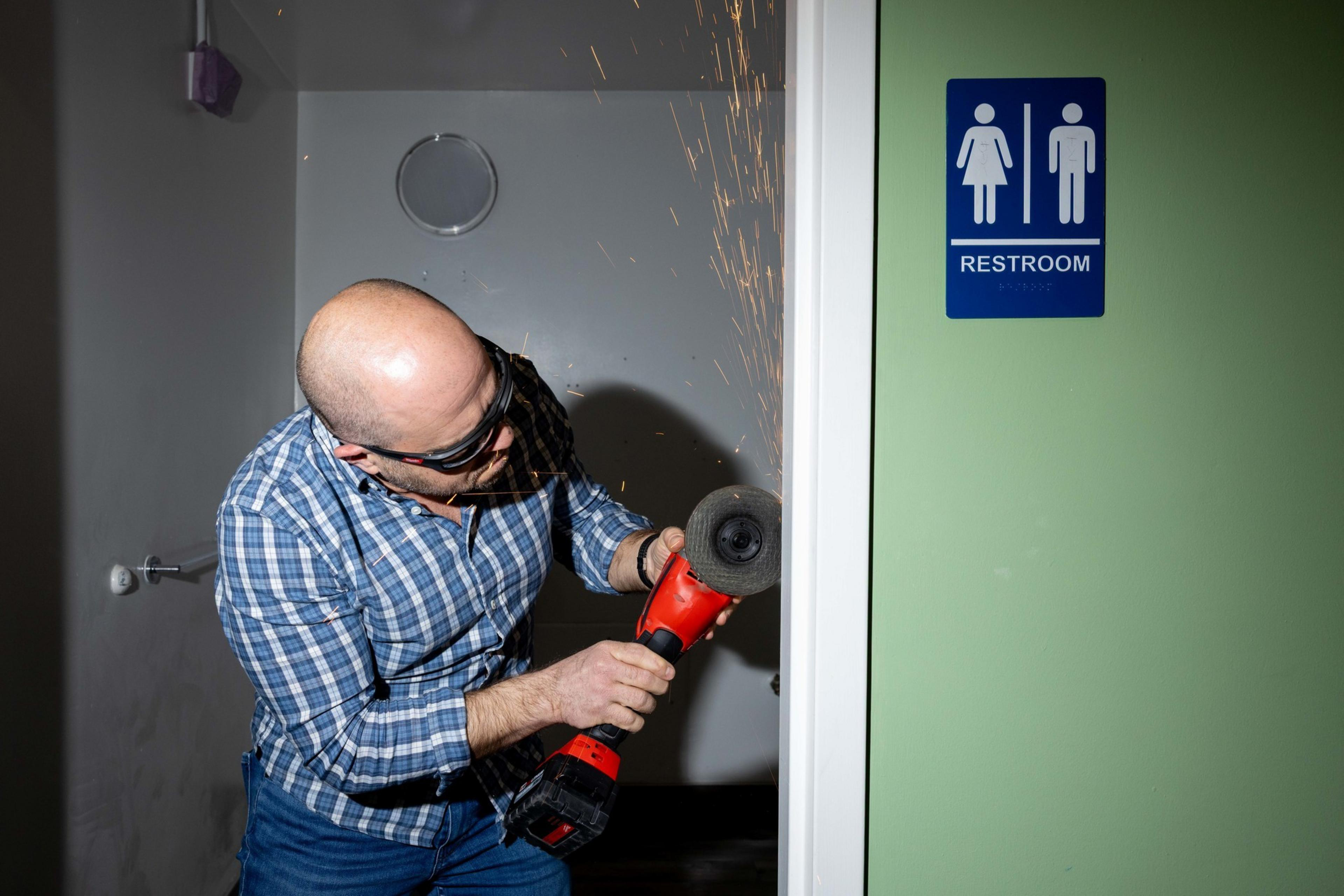 A person wearing goggles uses a red angle grinder on a restroom door frame, creating sparks. A restroom sign with male and female symbols is visible.