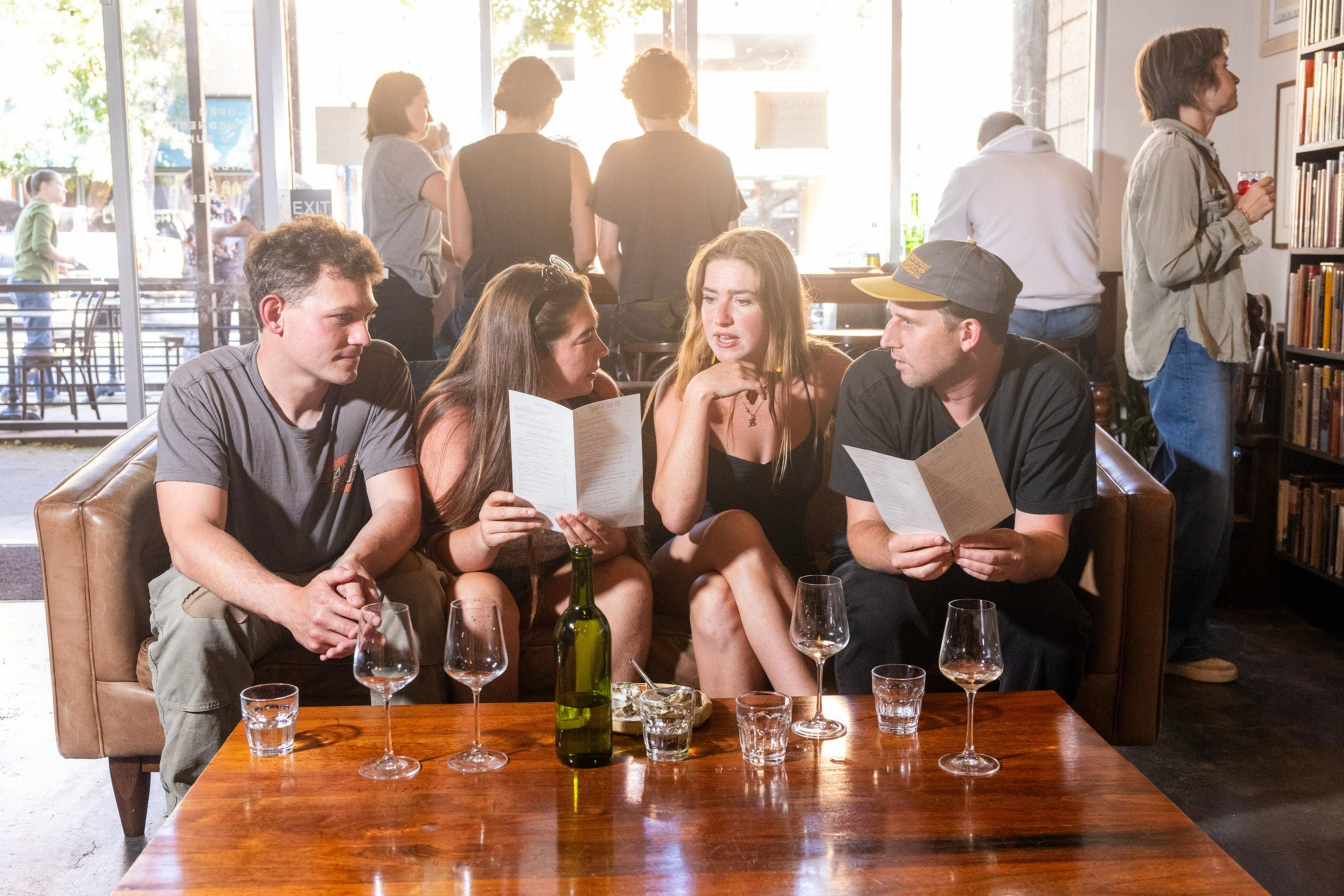 Four people sit on a couch in a lively café, holding menus. A wooden table with glasses and a bottle is in front of them. Other patrons stand and chat nearby.