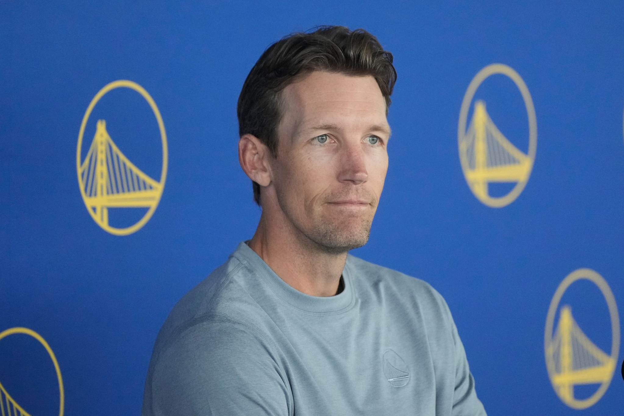 A man with short brown hair and blue eyes wears a gray shirt, sitting in front of a blue background featuring yellow Golden State Warriors logos.
