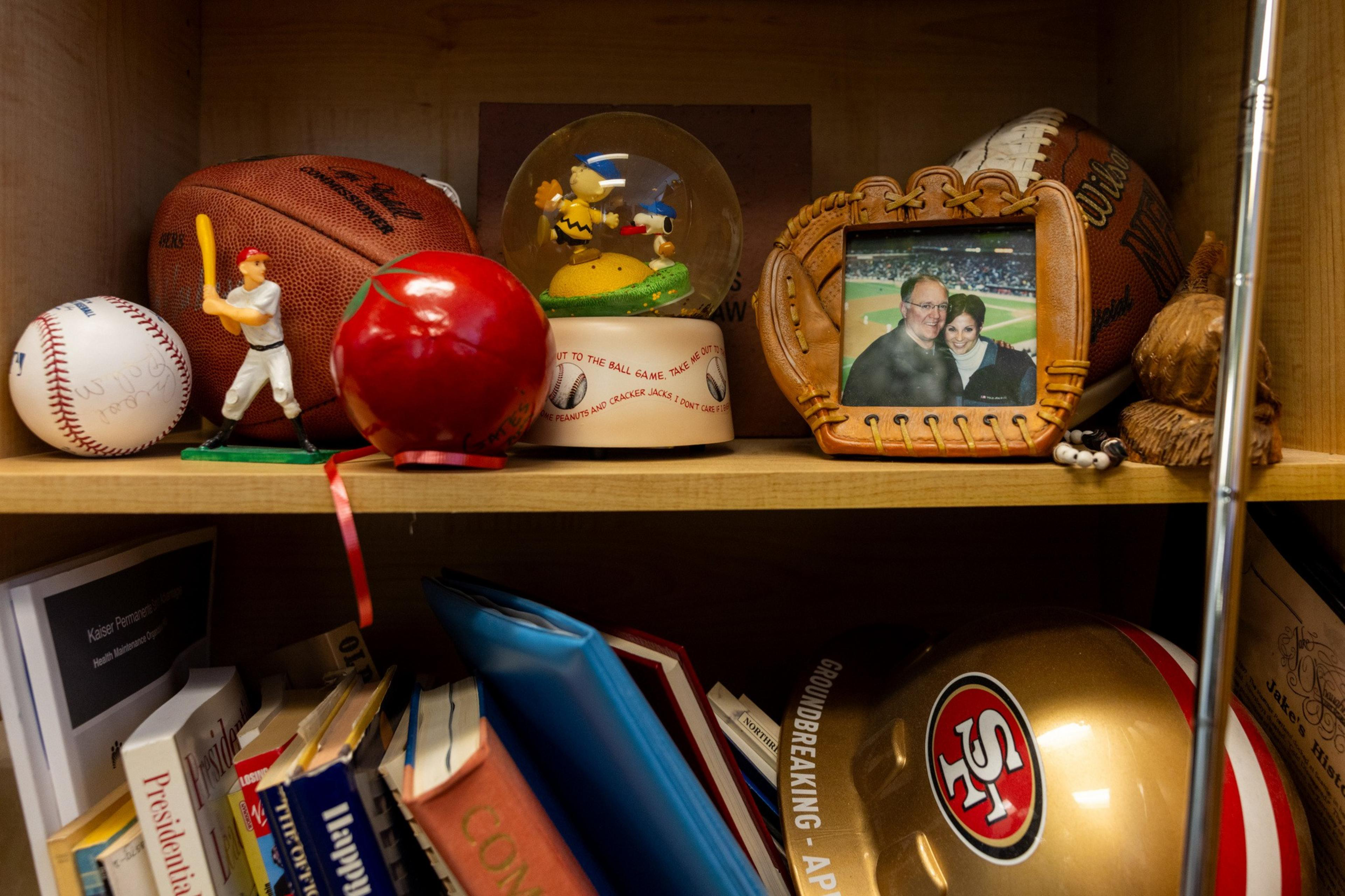 A shelf holds a basketball, baseball, figurine, snow globe, photo in a mitt frame, red apple, books, and a 49ers helmet. Other small objects are also present.