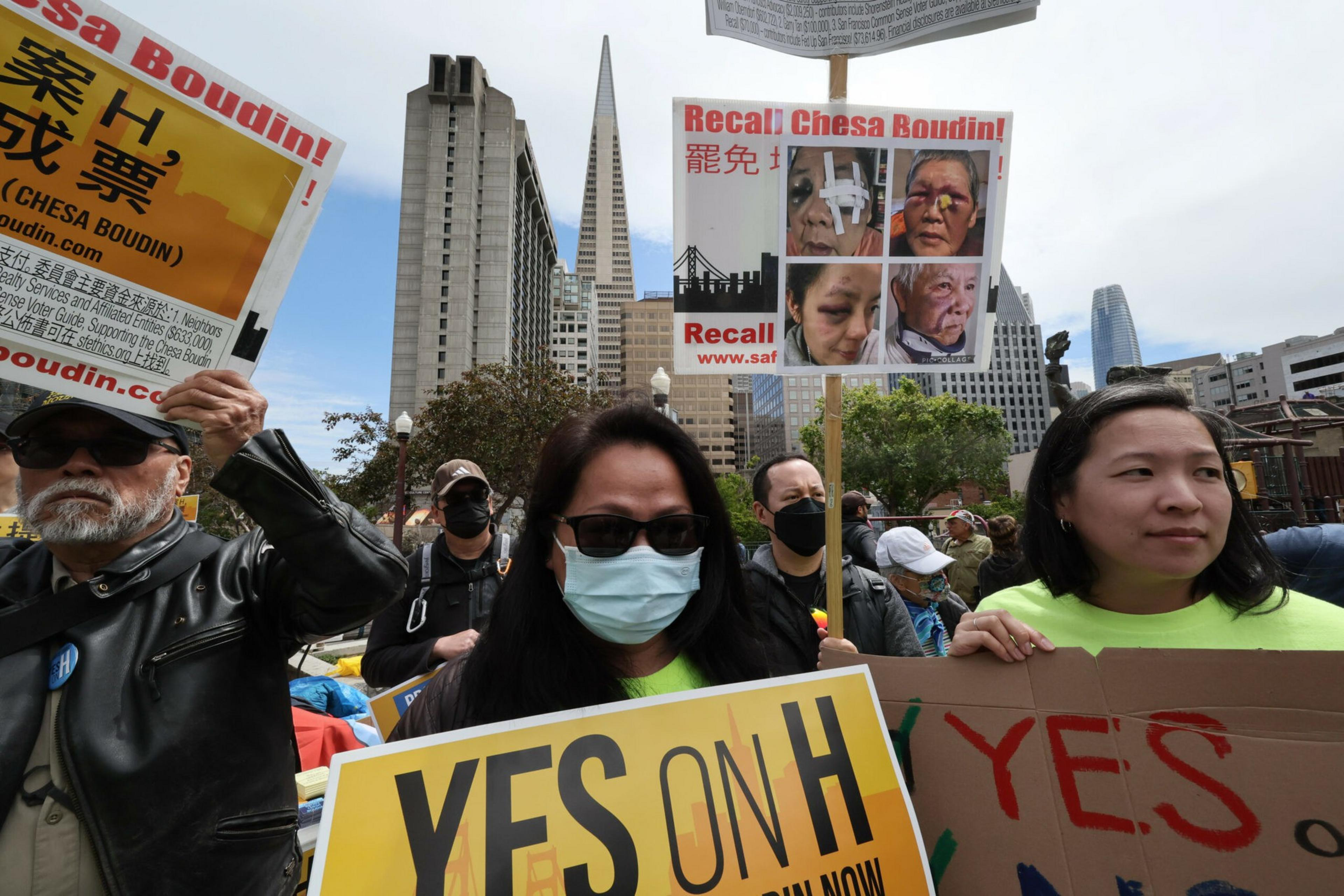 Protesters hold signs reading "YES ON H" and "Recall Chesa Boudin" in front of tall San Francisco buildings, with some wearing masks and sunglasses.