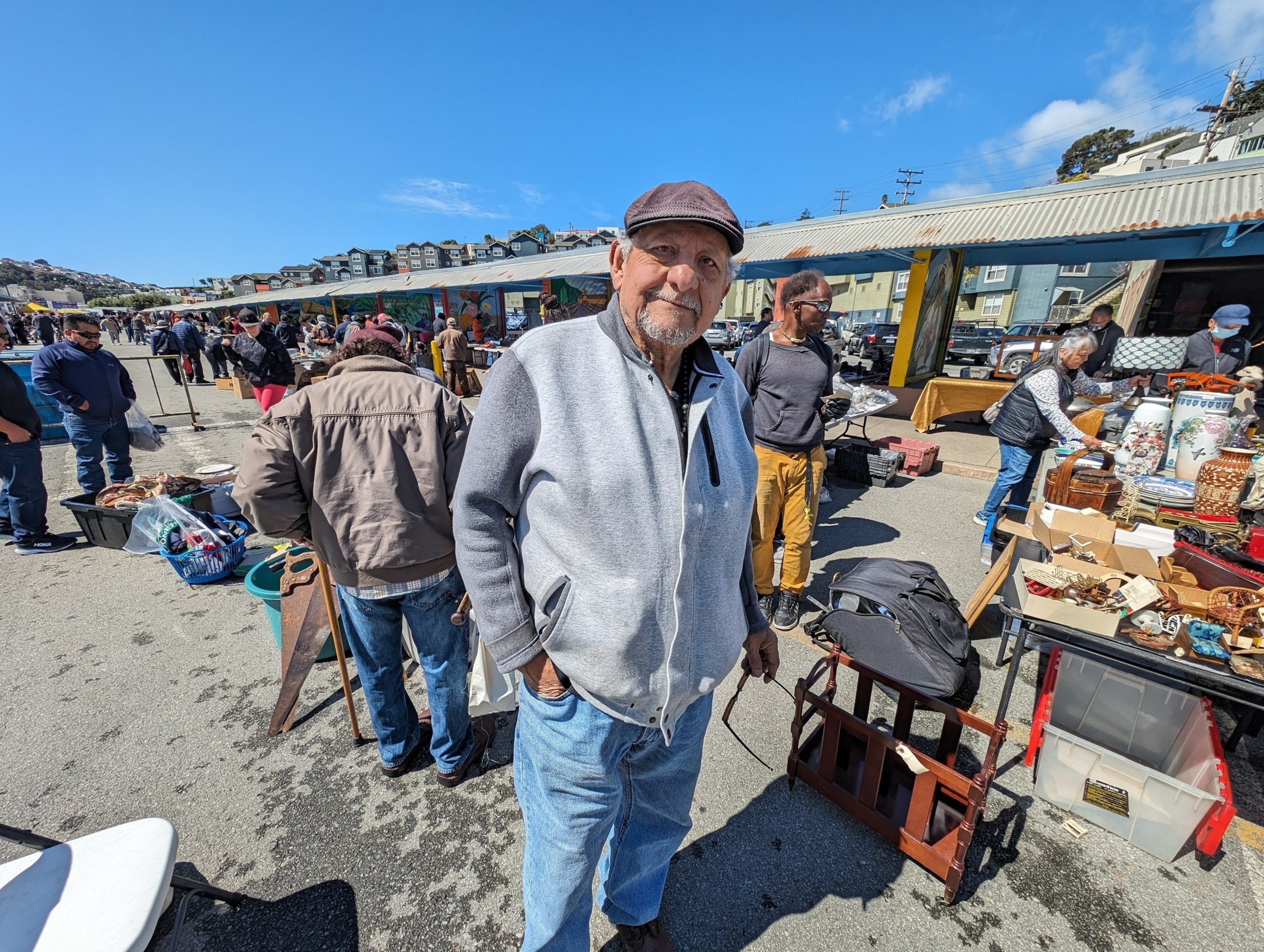 A goateed man wearing a gray fleece jacket, brown cap and jeans stands in sunshine