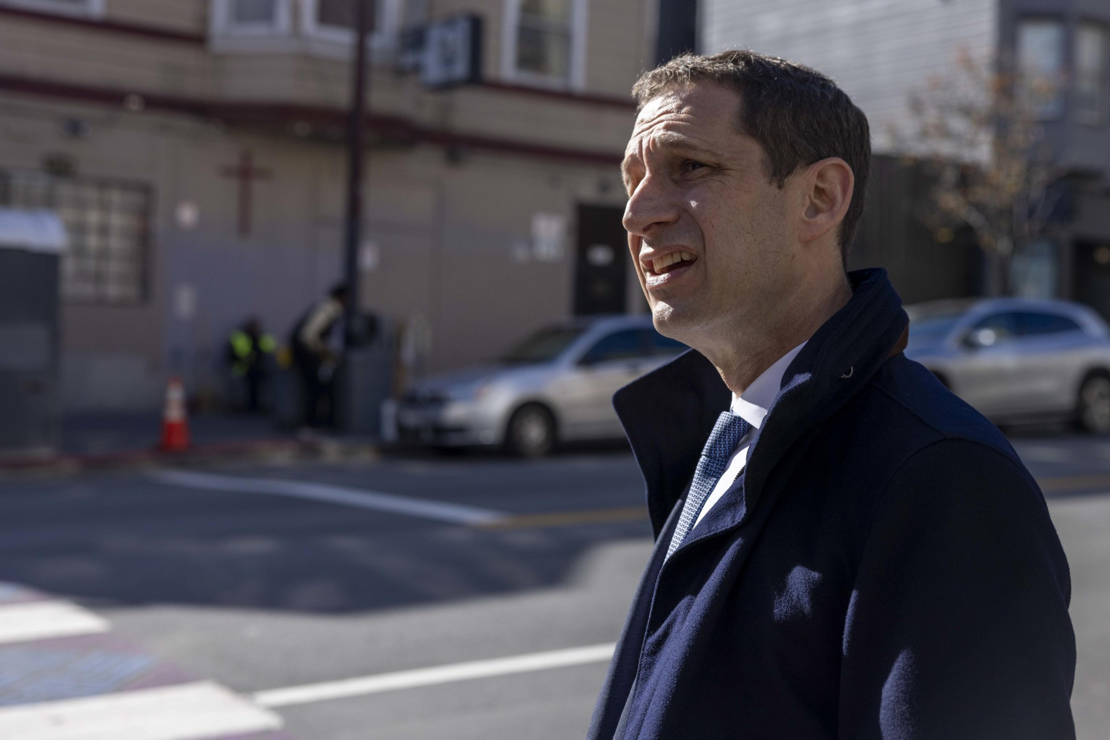 A man in a dark coat stands on a sunny street, speaking. In the background, there's a building with a cross, a car, and people near a traffic cone.
