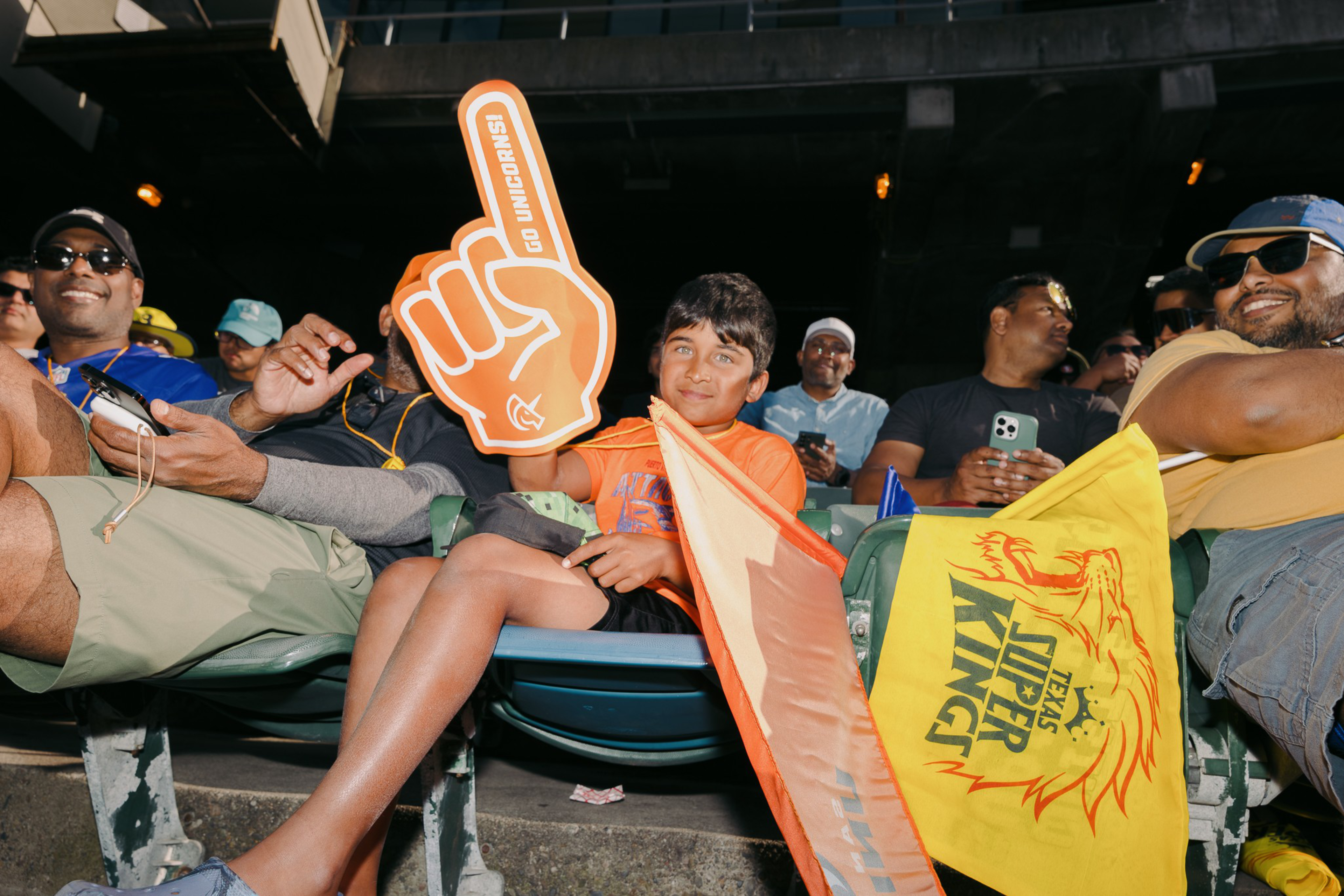 A young boy at a stadium holds an orange foam finger and flags. People around him are seated, some using smartphones. One flag reads &quot;Texas Super Kings.&quot;
